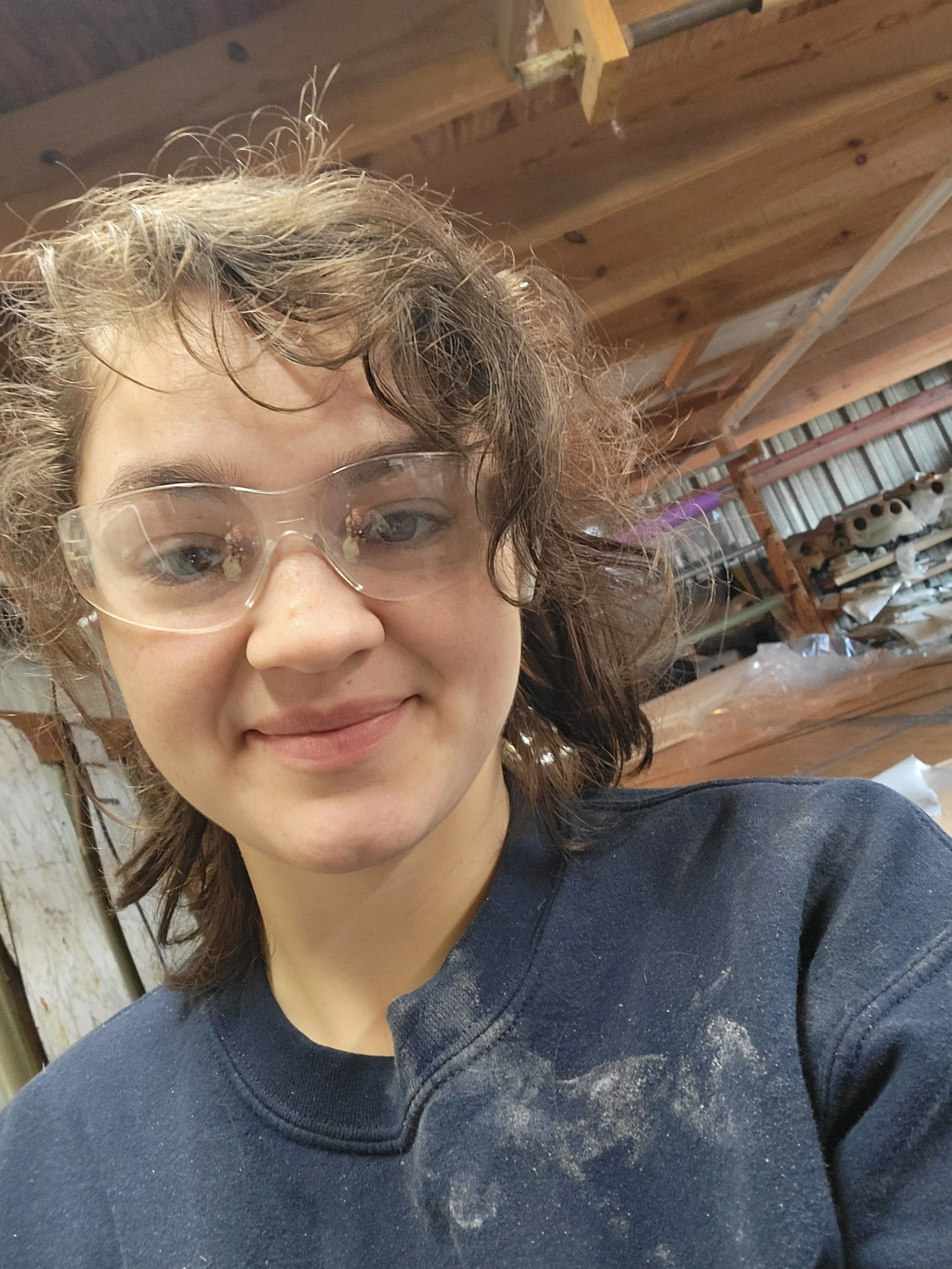Close-up of a woman with curly hair wearing protective glasses and a black shirt, standing in a workshop with wooden shelves and tools in the background.