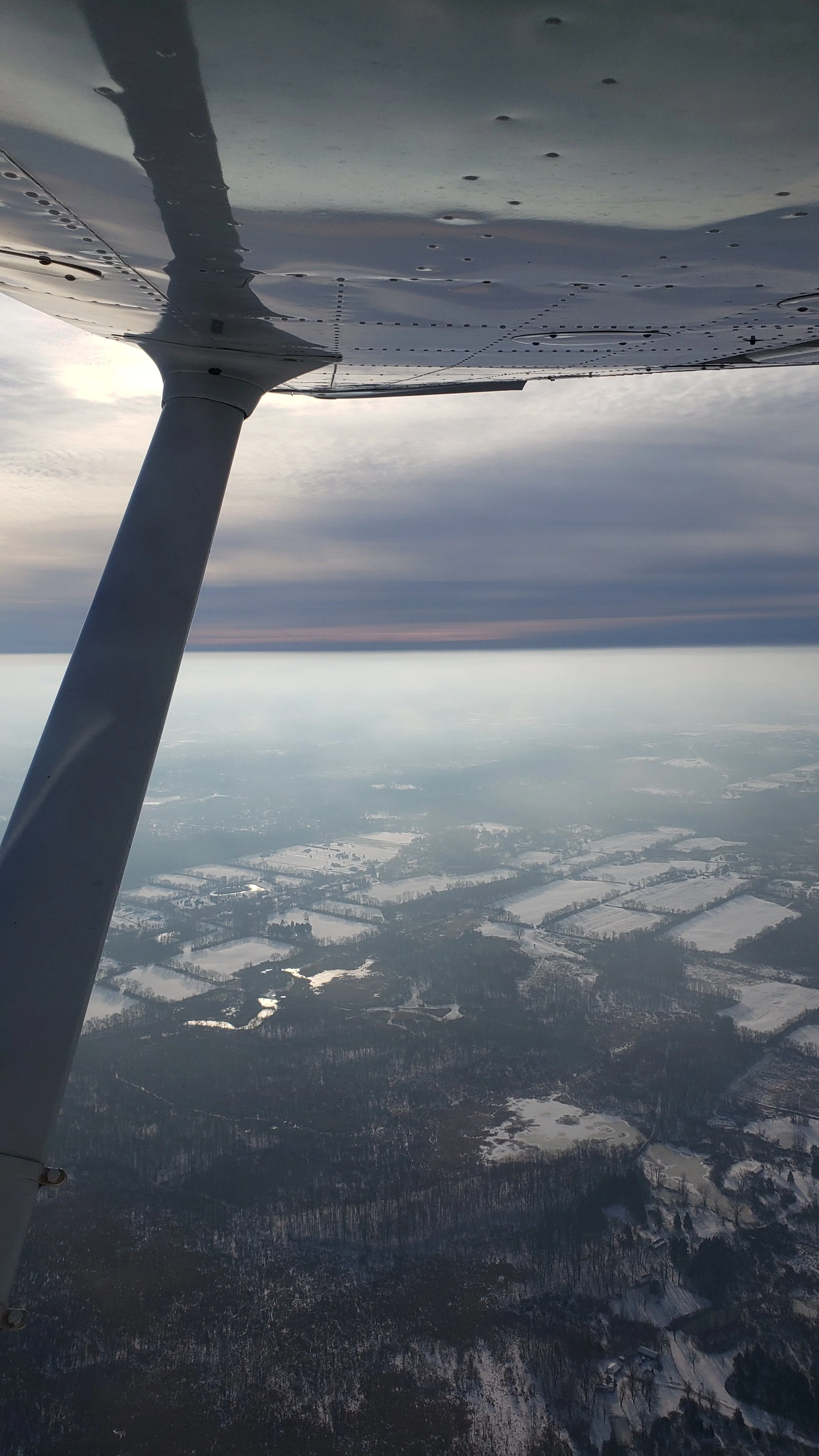 Aerial view of snow-covered farmland and forests taken from a small airplane, with a support strut visible in the foreground.