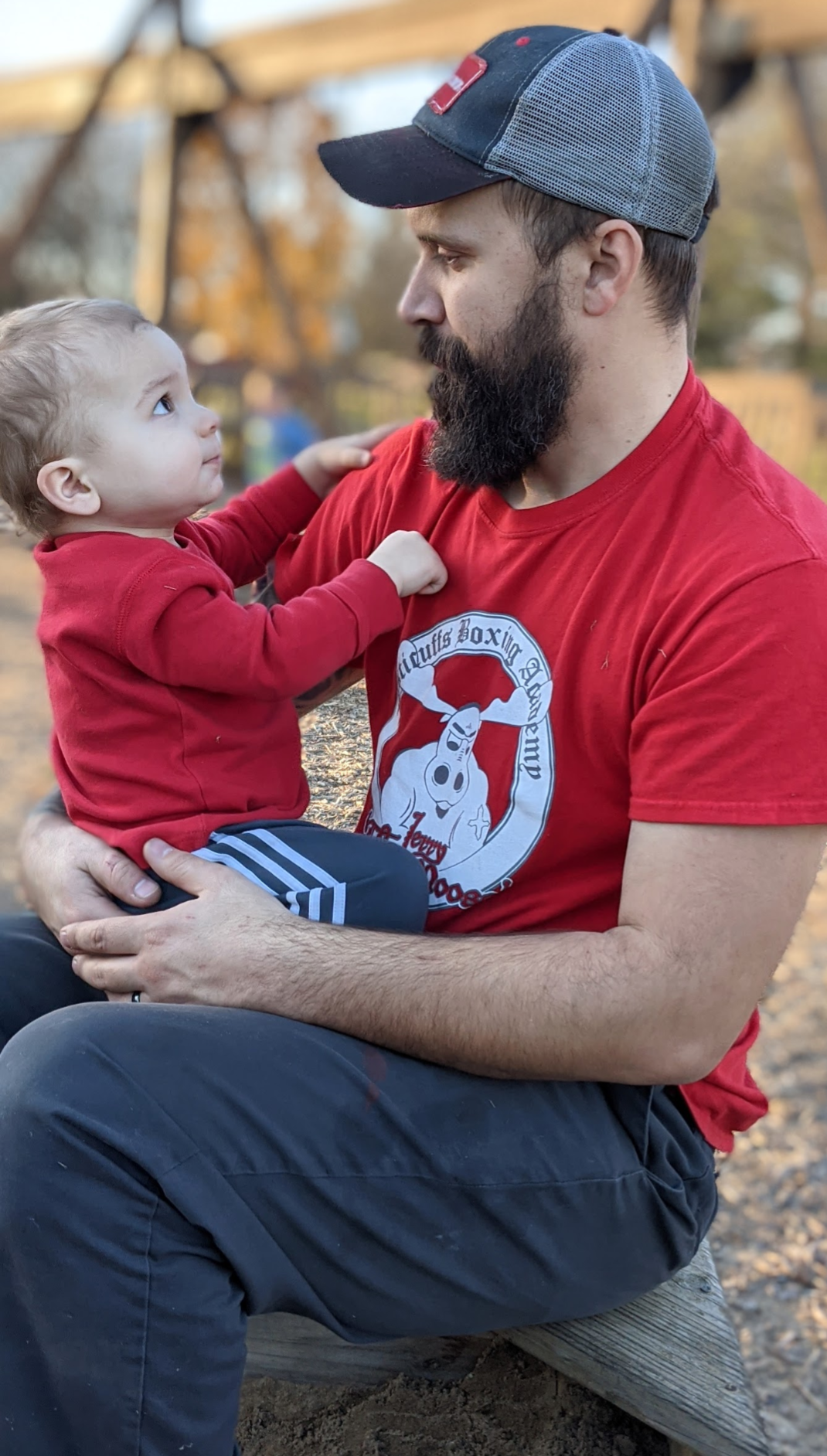 A man with a beard and a cap holding a young child in a red hoodie and striped pants, sitting outdoors on a park bench during autumn.