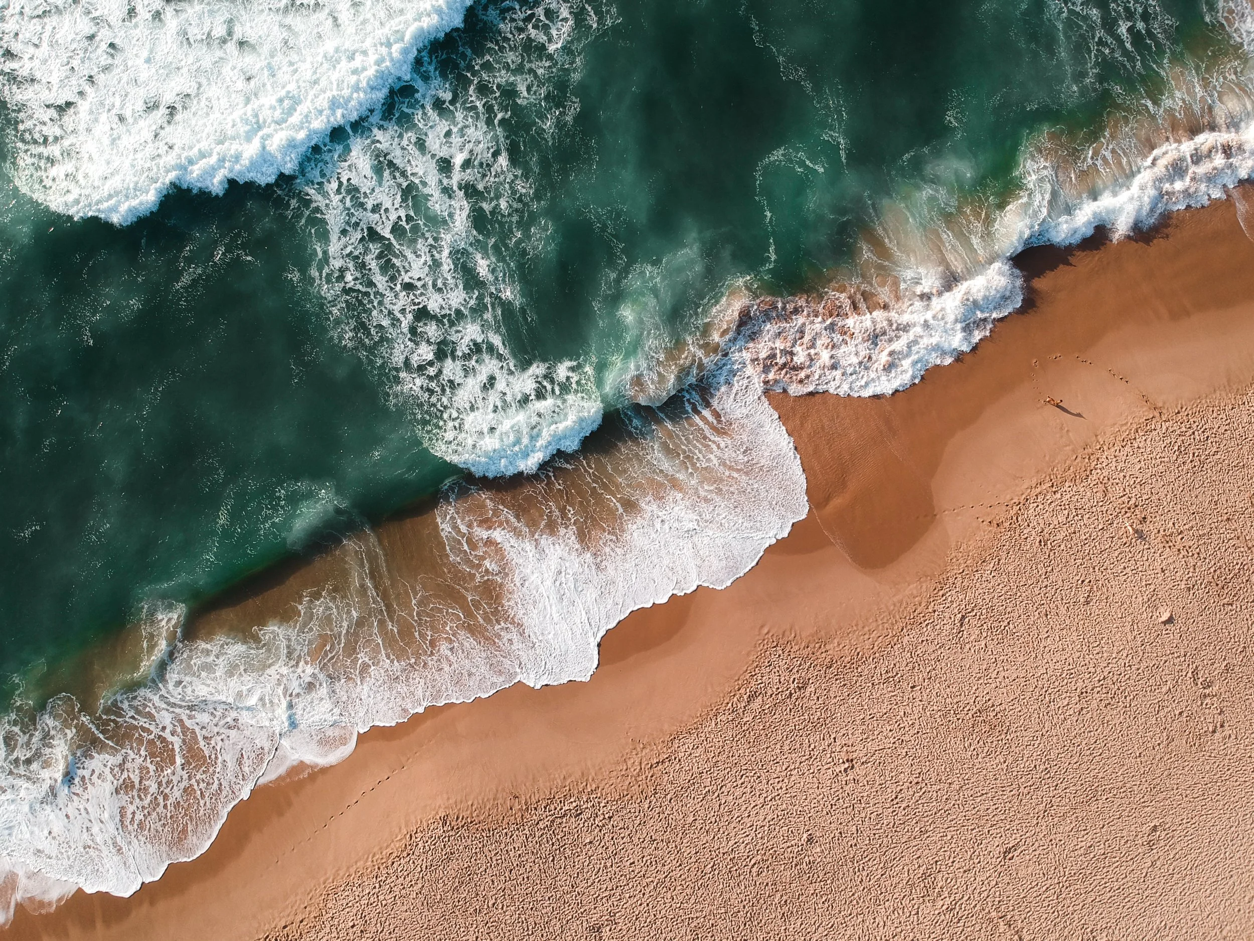 The image shows an aerial view of the ocean with white waves crashing onto the sandy beach, and a person walking along the shoreline.