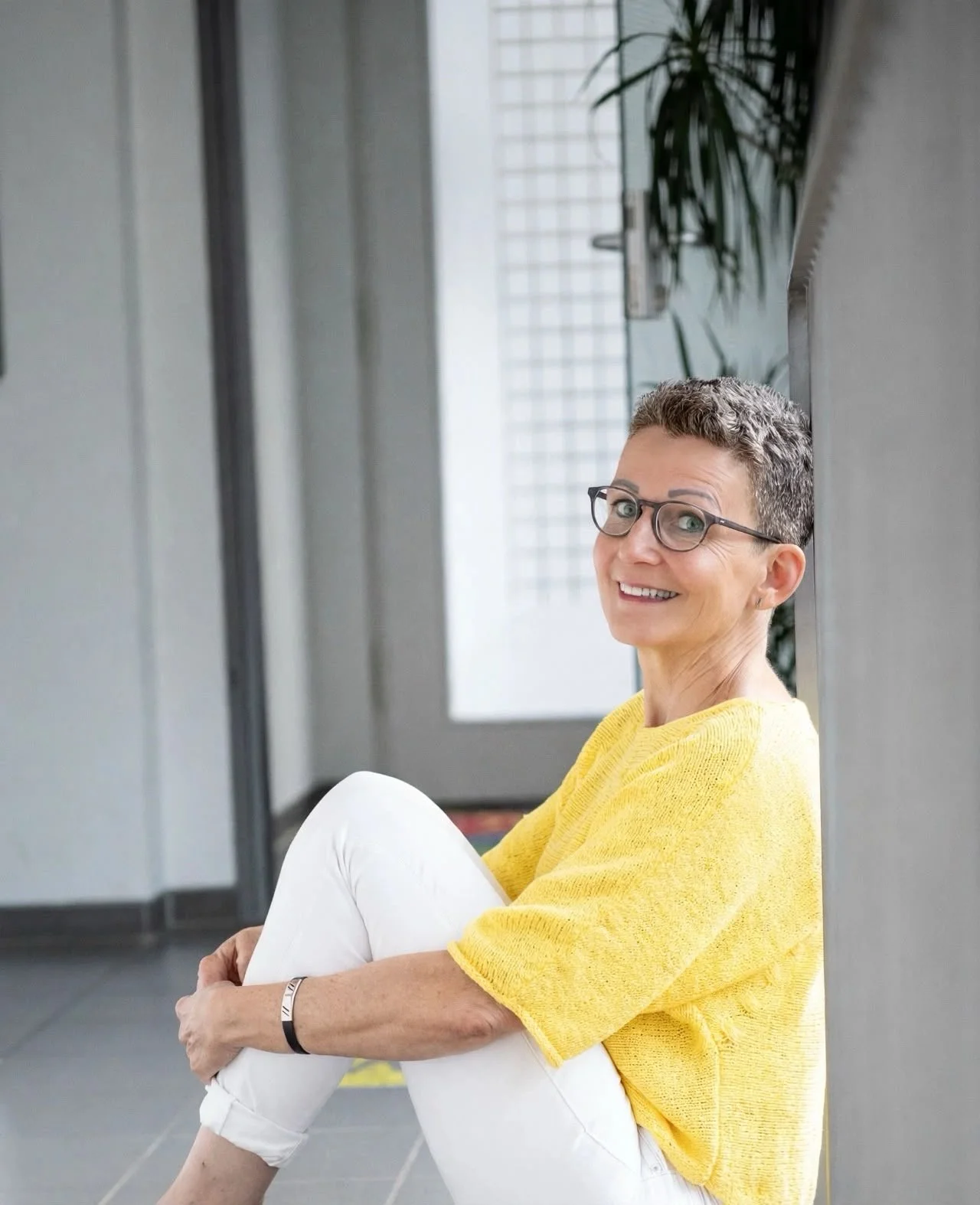 Lächelnde Frau mit Brille und gelbem Pullover sitzt entspannt auf dem Boden neben einem Fenster.