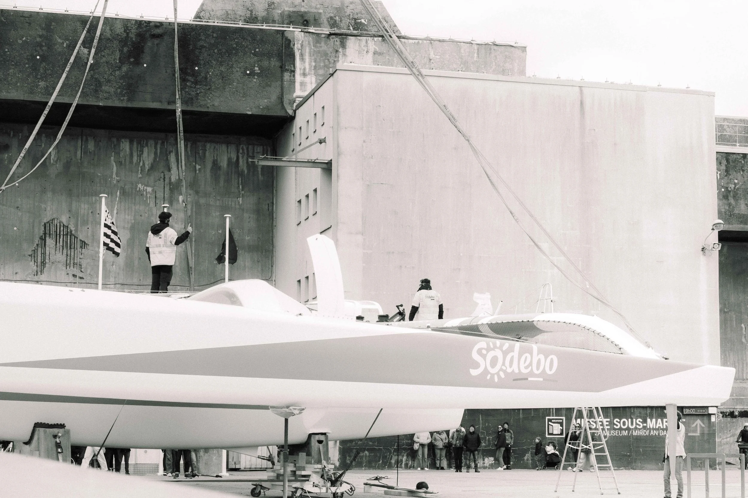 Une maquette de voilier électrique à l'extérieur, avec des personnes en arrière-plan près du bâtiment.