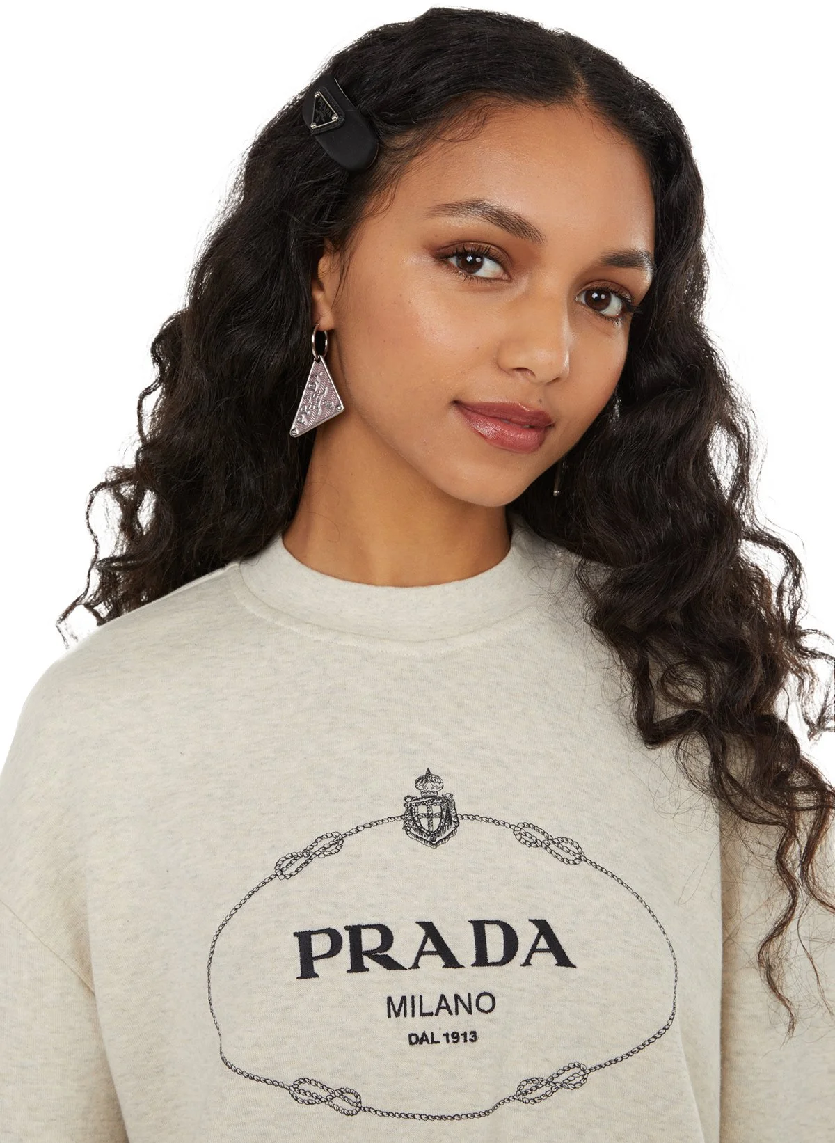Jeune femme portant un sweat-shirt beige à logo Prada et une épingle à cheveux noire, avec boucle d'oreille en forme de triangle en métal.