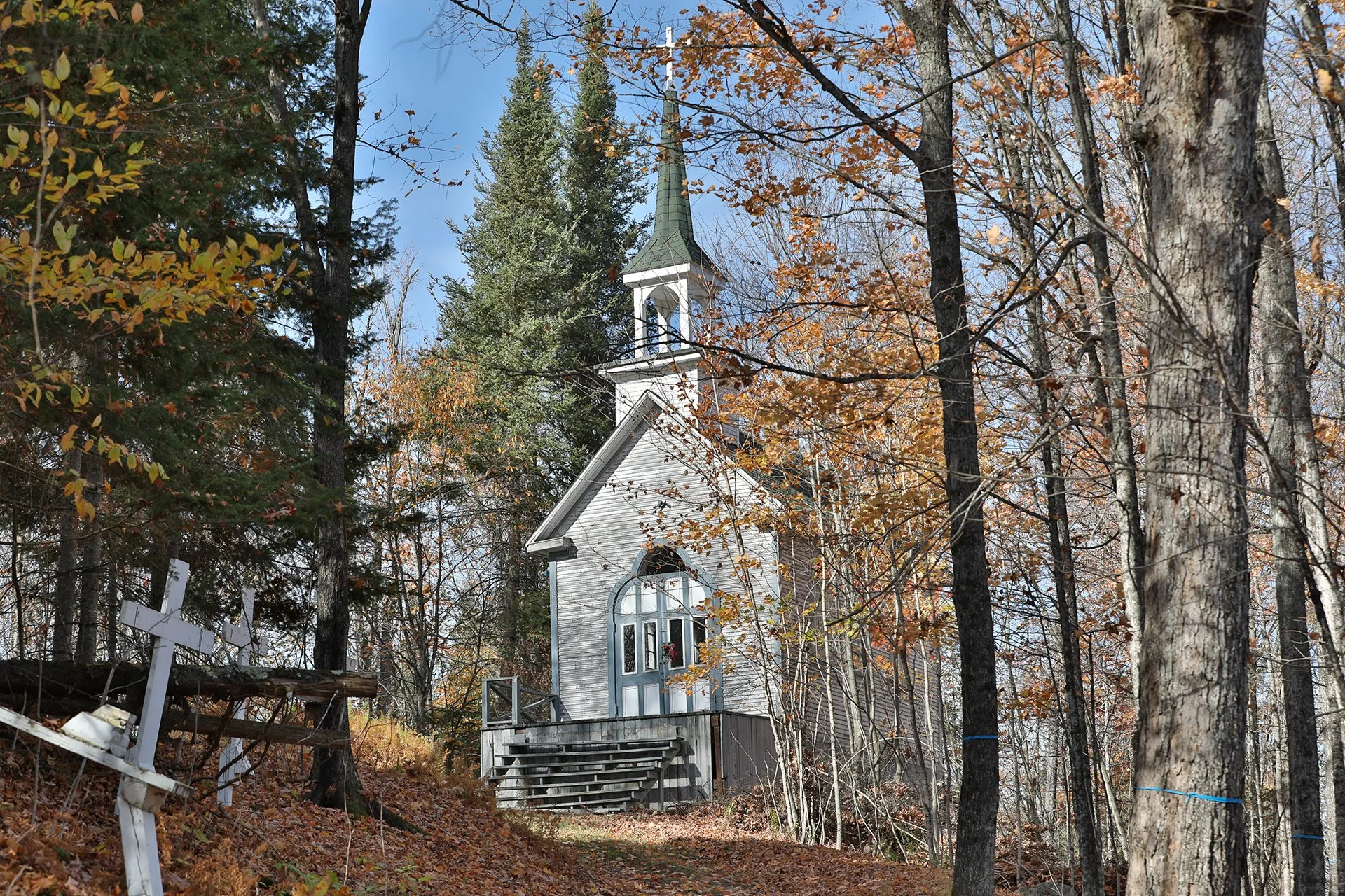 Une petite église en bois gris dans une forêt en automne, entourée de arbres aux feuilles tombées, avec un ciel bleu clair.