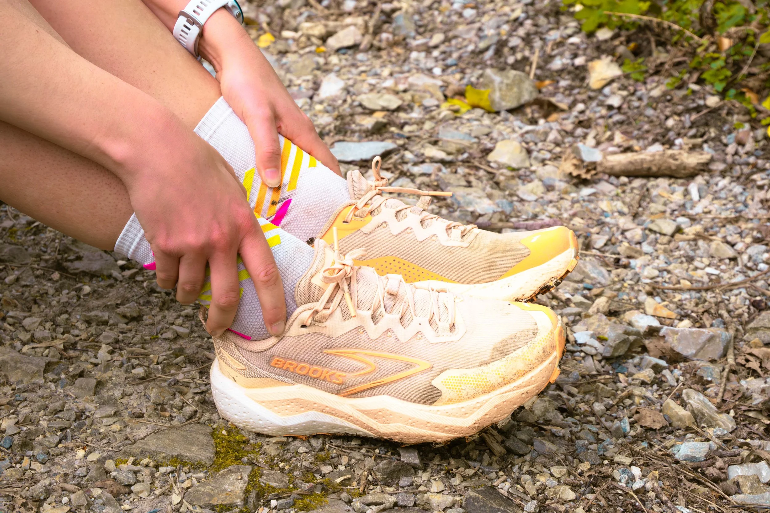 Personne attachant des chaussures de course beige et jaune sur un sentier rocheux, portant des chaussettes blanches et une montre blanche.