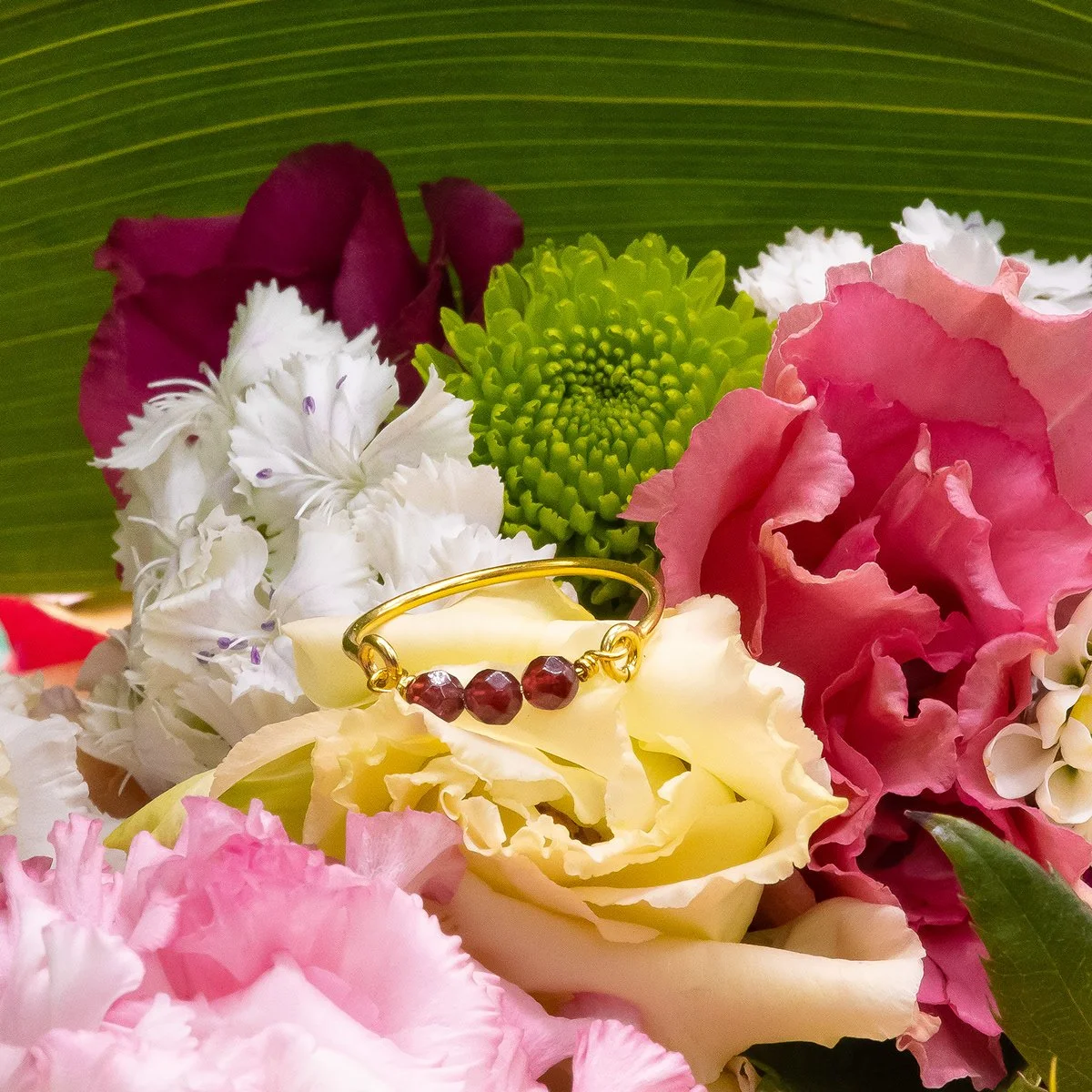 Collier en or avec trois pierres rouges, posé sur un bouquet de fleurs de différentes couleurs, y compris blanc, rose, violet et vert, avec un fond de feuille verte