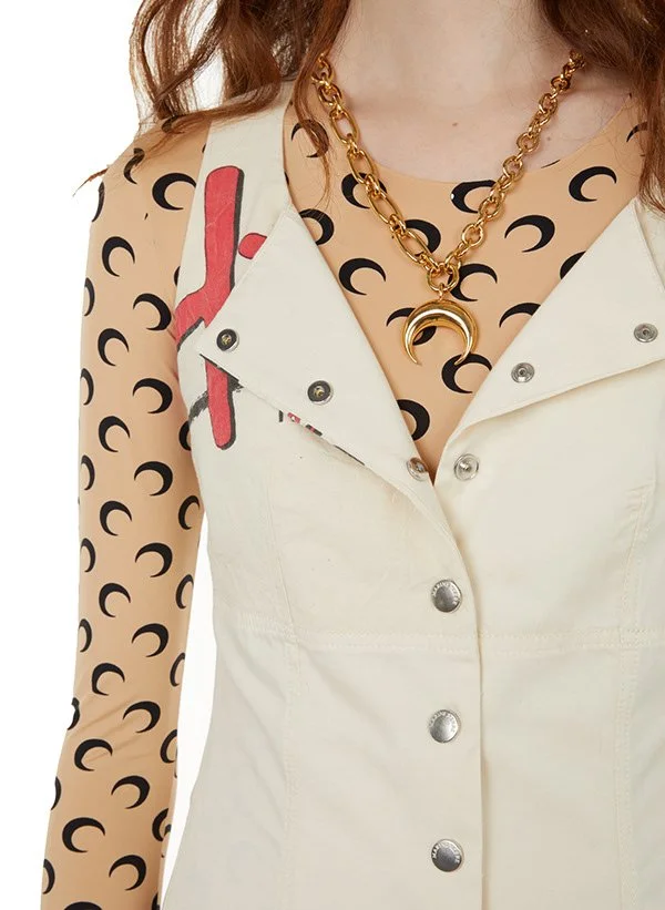 Une femme porte une veste blanche avec un motif de petits croissants noirs et une chemise en mesh beige avec des motifs rouges et noirs. Elle porte un collier épais en or avec un pendentif croissant doré.