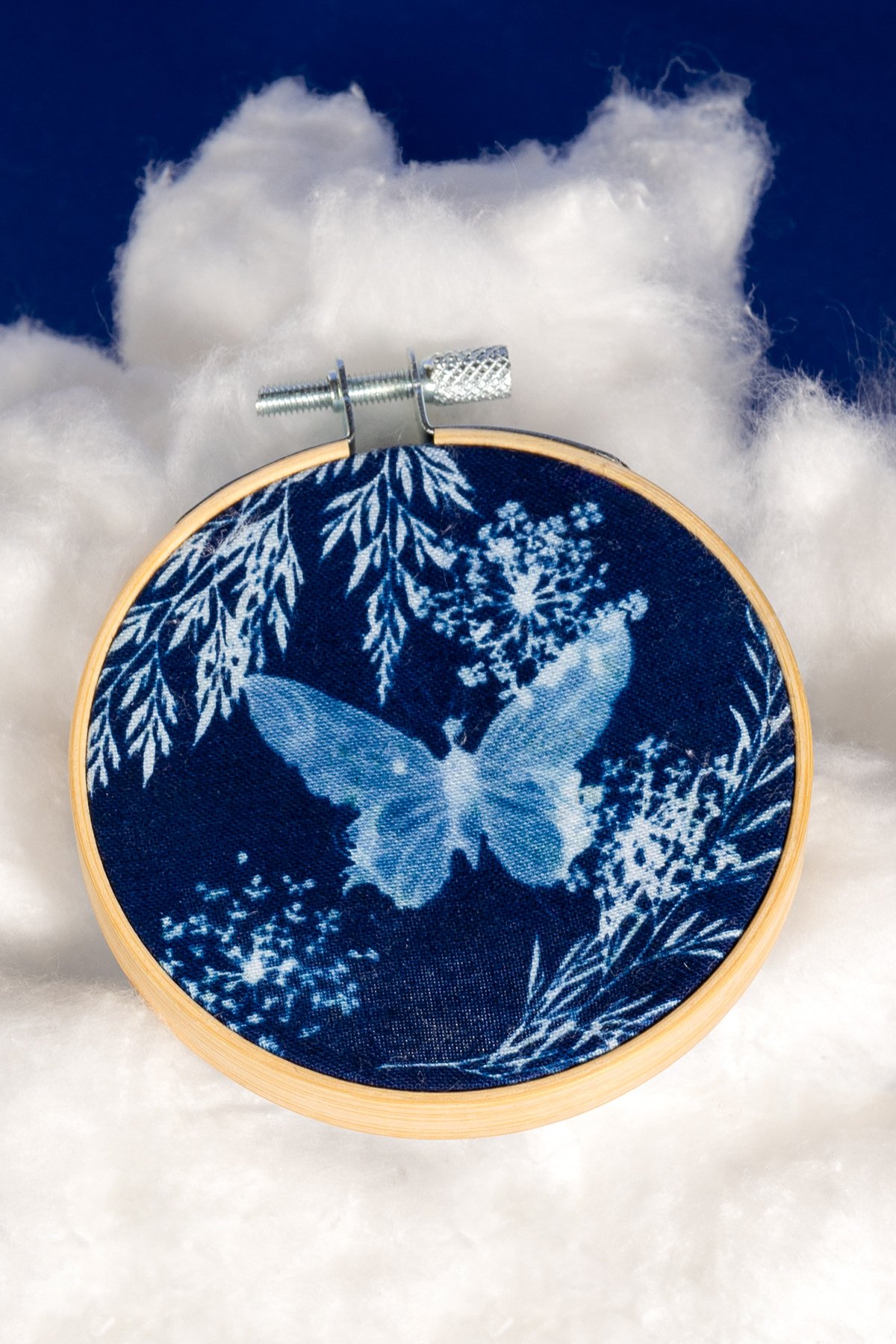 Un cercle en bois avec un tissu bleu représentant un papillon blanc et des motifs floraux, placé sur une surface de coton ou de nuages blancs, face à un fond bleu.