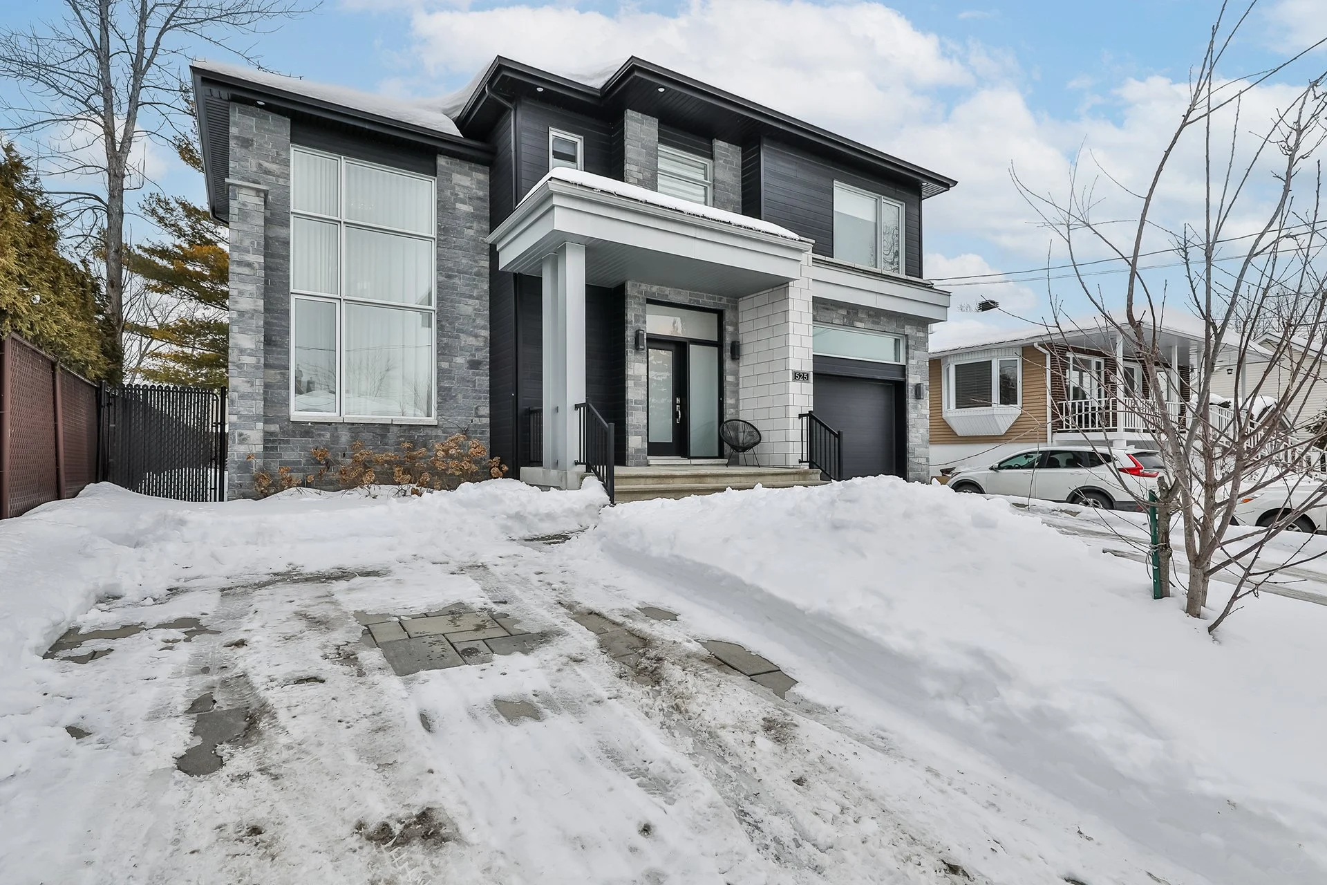 Maison moderne à deux étages avec façade en pierre grise, fenêtres grandes et garage, entourée de neige, avec un ciel partiellement nuageux.