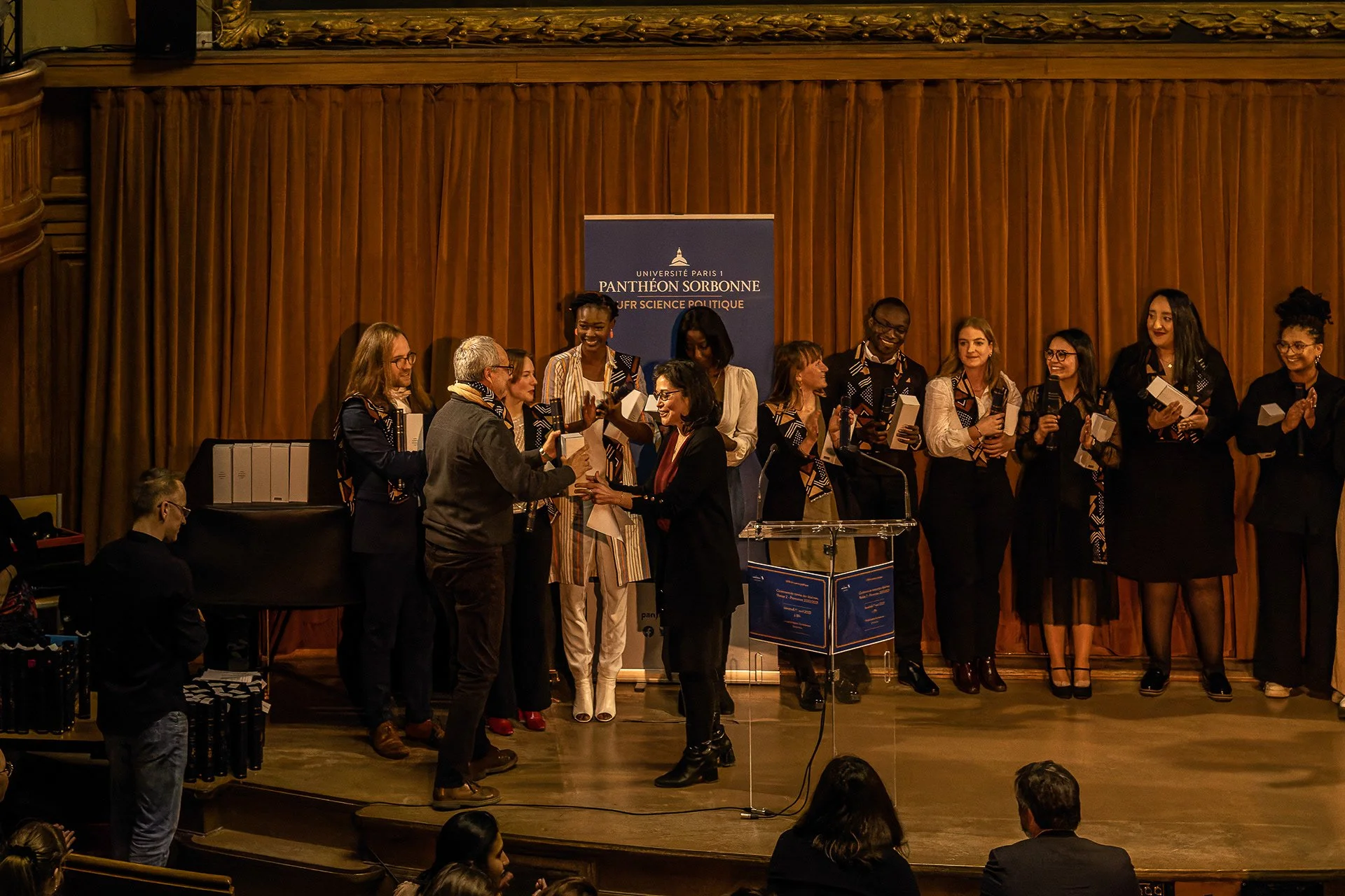 Cérémonie académique avec remise de trophée ou diplôme sur scène, plusieurs personnes applaudissent, un fond avec une bannière de l'Université Paris 1 Panthéon Sorbonne.