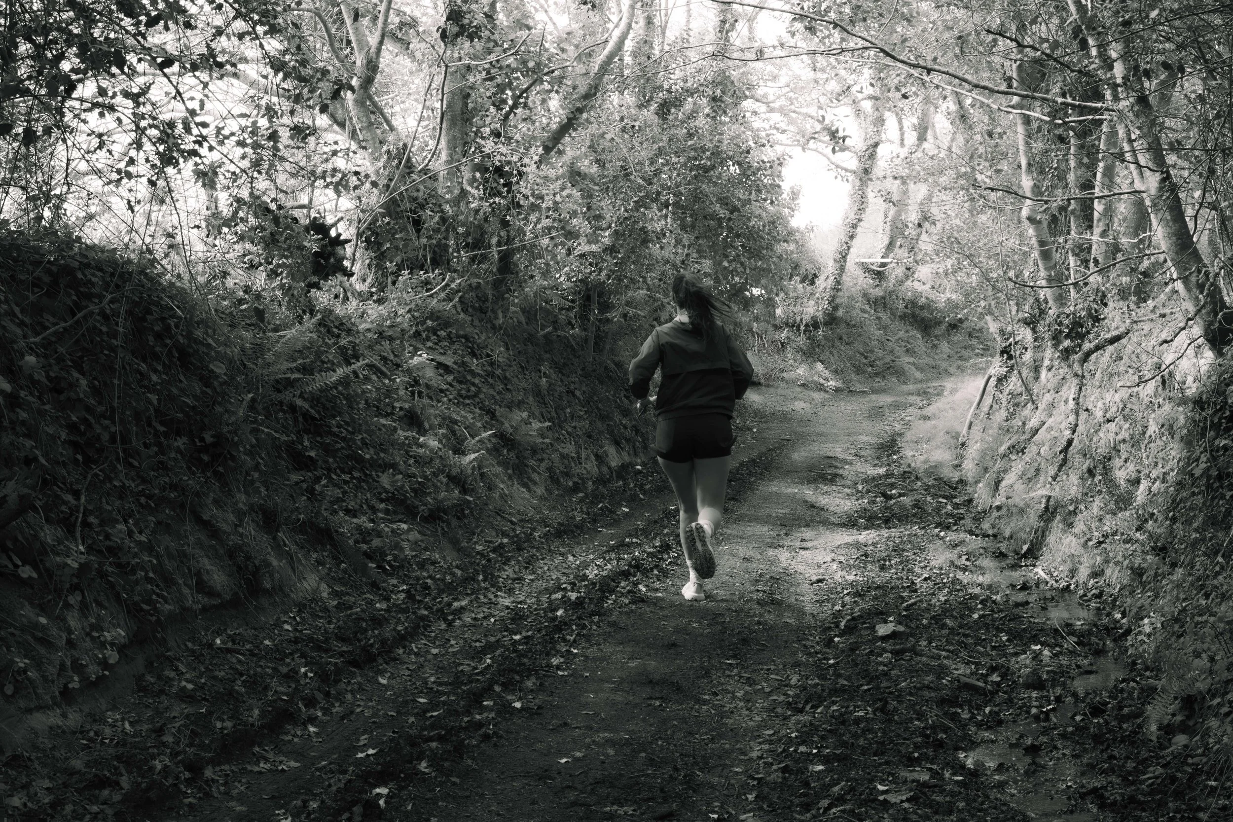 Une femme court sur un sentier de forêt bordé d'arbres et de végétation dense, vue de dos, en noir et blanc.