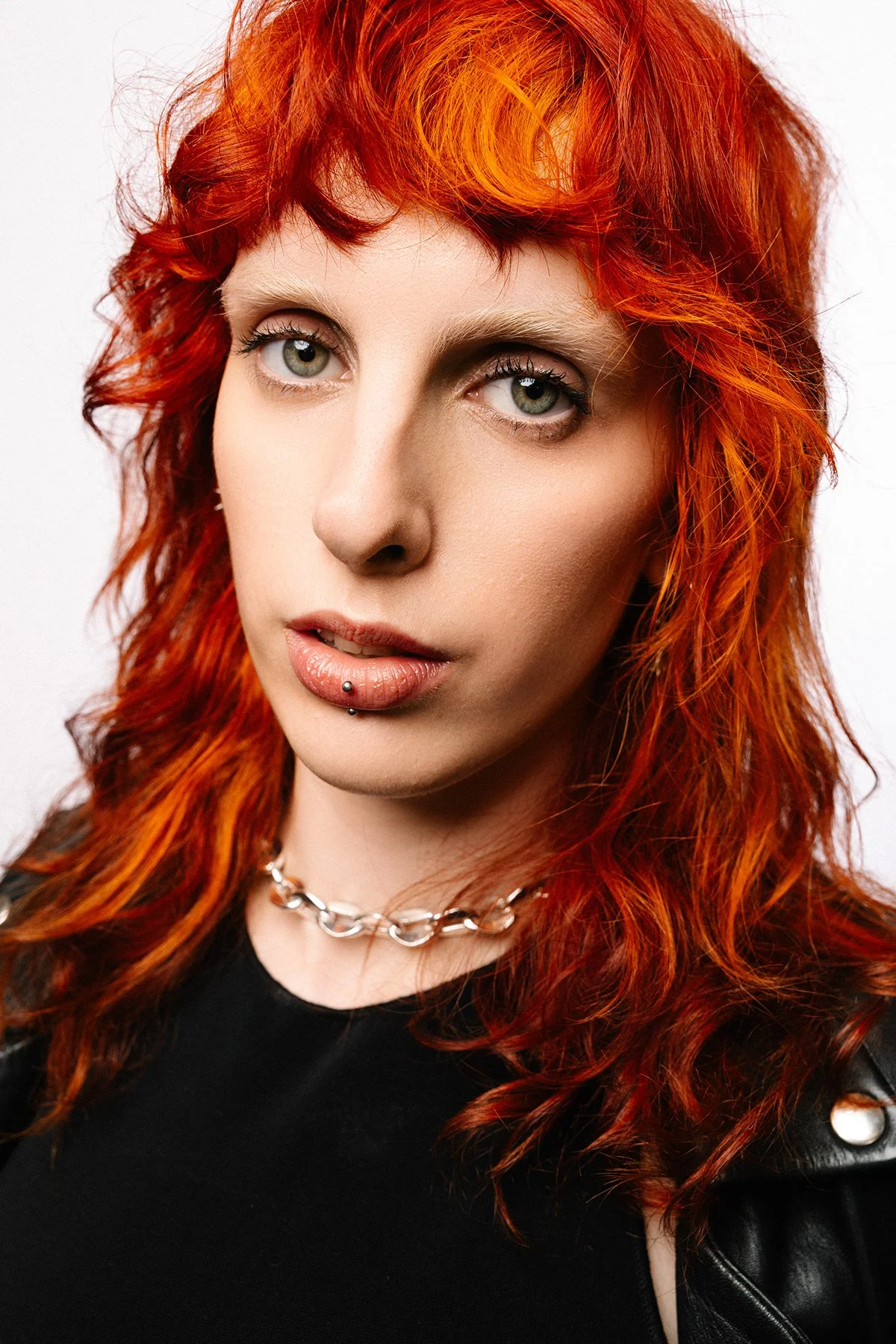 Portrait d'une femme avec des cheveux roux ondulés, portant un collier en chaîne et un piercing à la lèvre, portant un vêtement noir.
