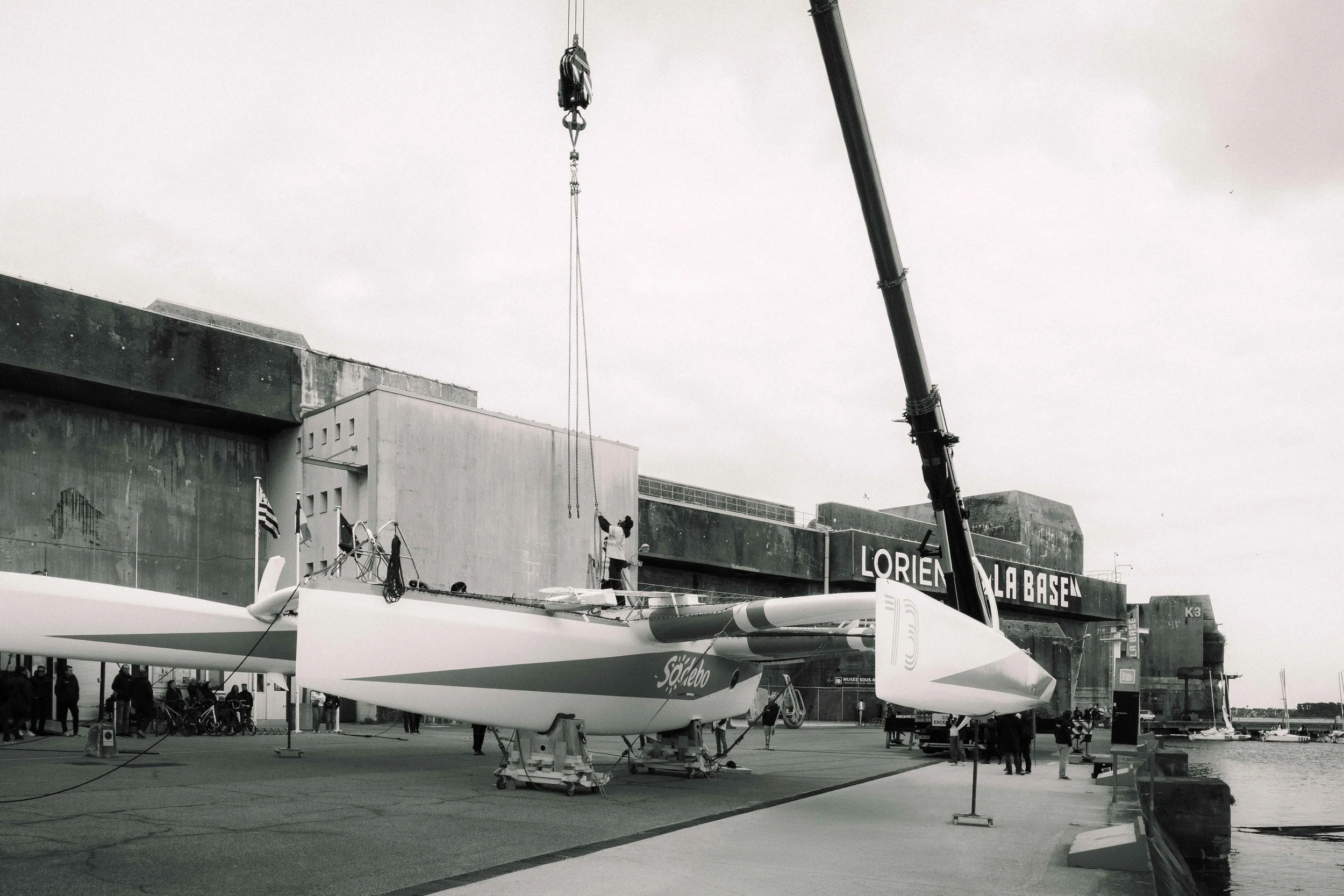 Une voile de bateau à voile stationnée devant un bâtiment en béton, avec des personnes autour et un abri de bateau à proximité, au bord de l'eau, en noir et blanc.