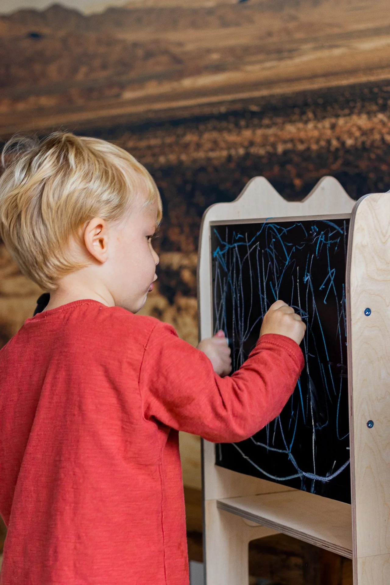 Jeune enfant blond dessine sur un tableau noir avec des traits de craie blanche.
