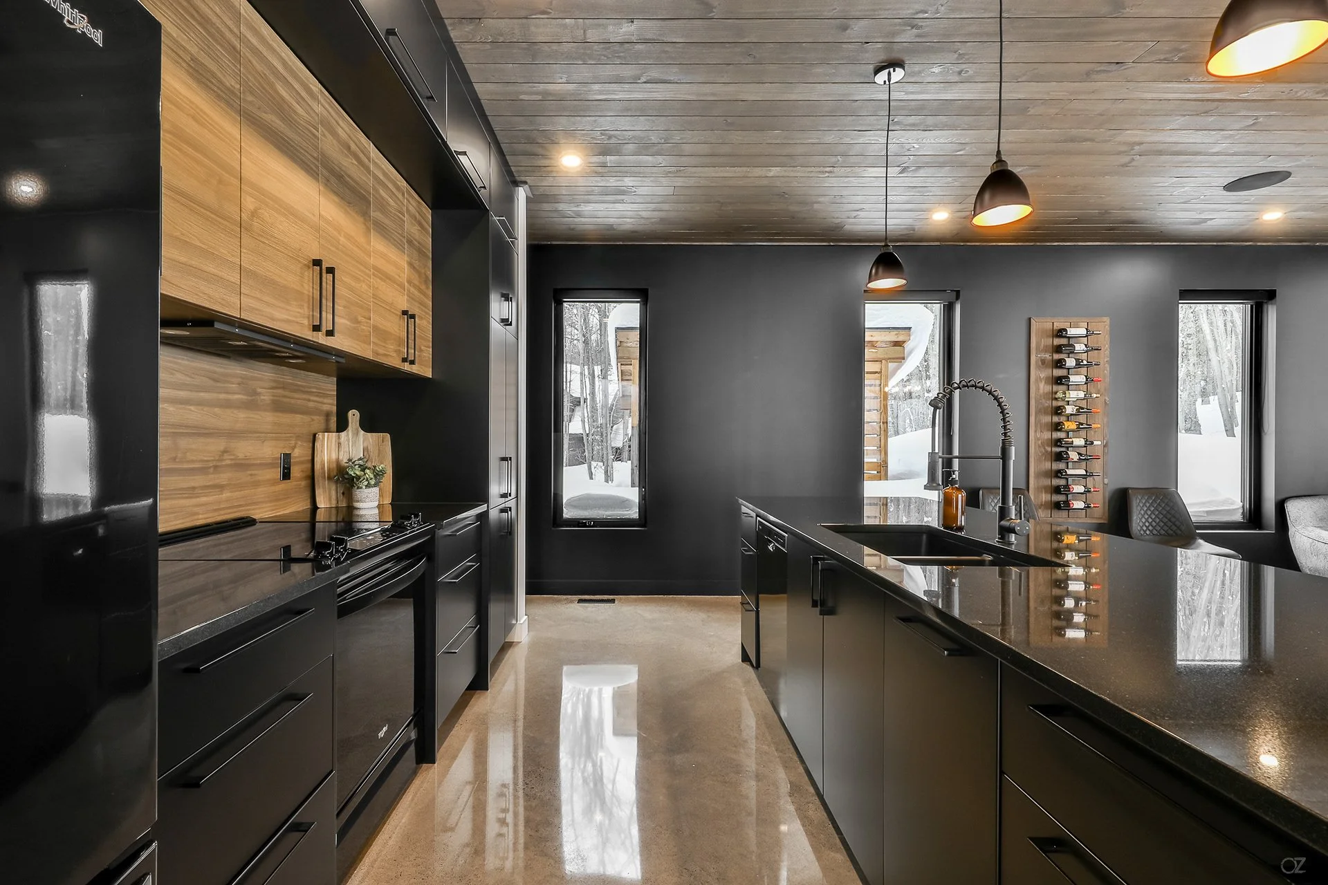 Cuisine moderne avec armoires noires, plan de travail en granite noir, murs gris foncé, et plafond en bois brut. Éclairage suspendu noir, vinothèque contre le mur, fenêtres donnant sur un espace enneigé.