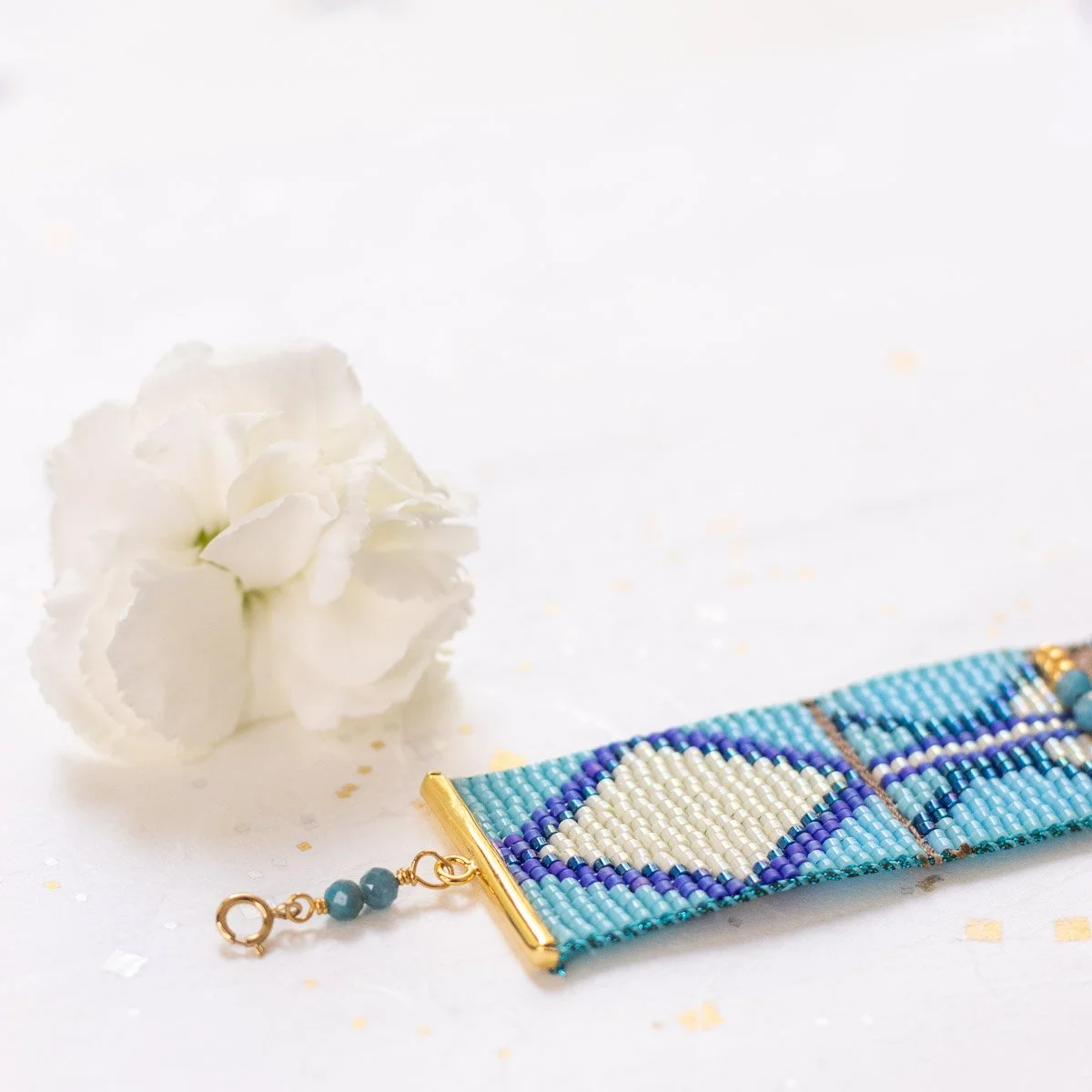 Bracelet en perles bleu et blanc avec motif de coeur, posé à côté d'une fleur blanche sur un fond blanc avec des reflets dorés.