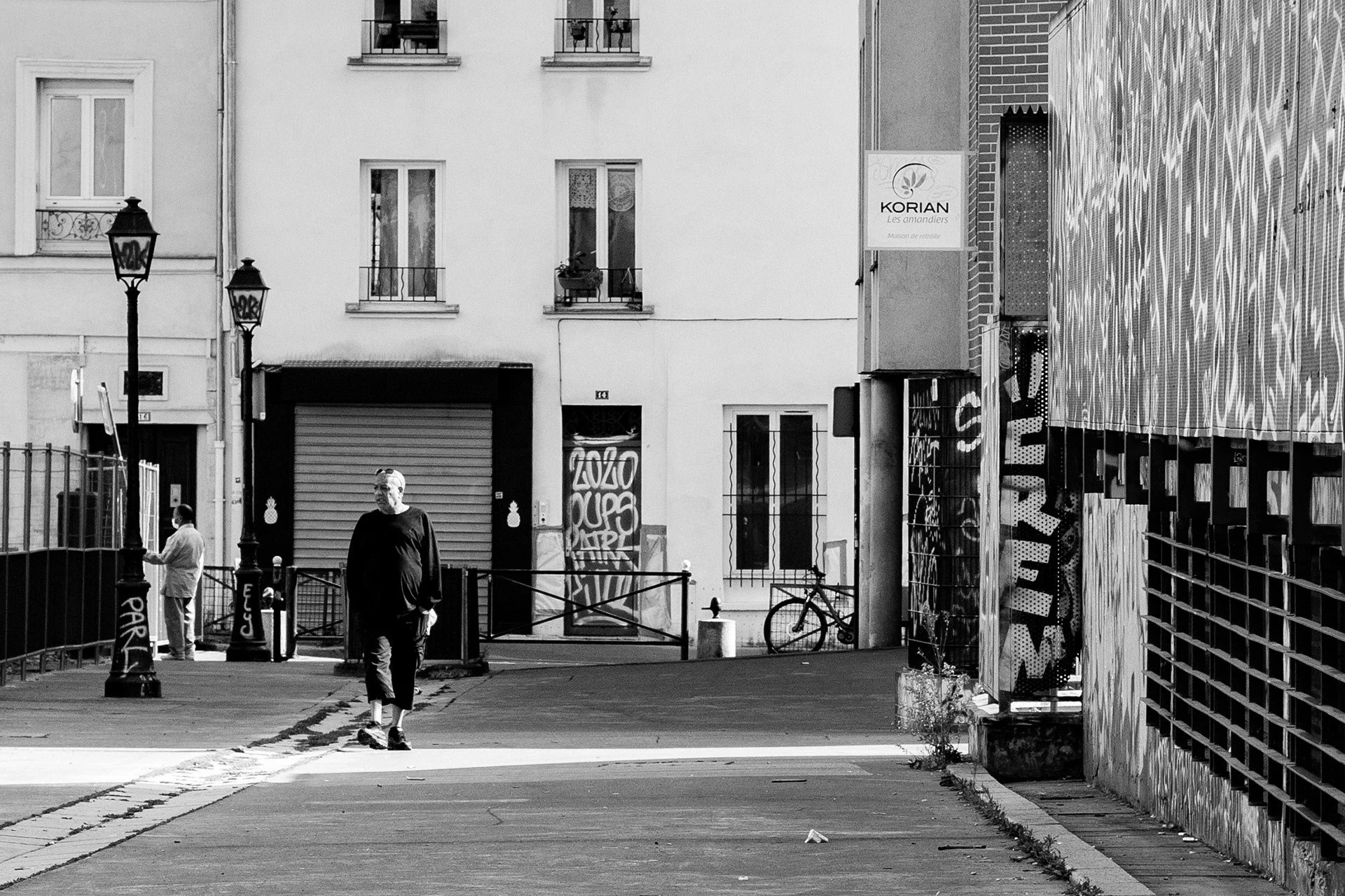 Une rue urbaine en noir et blanc avec deux personnes, une homme marchant vers l'avant et une autre au téléphone, des lampadaires, des graffitis sur les murs, et un vélo stationné.