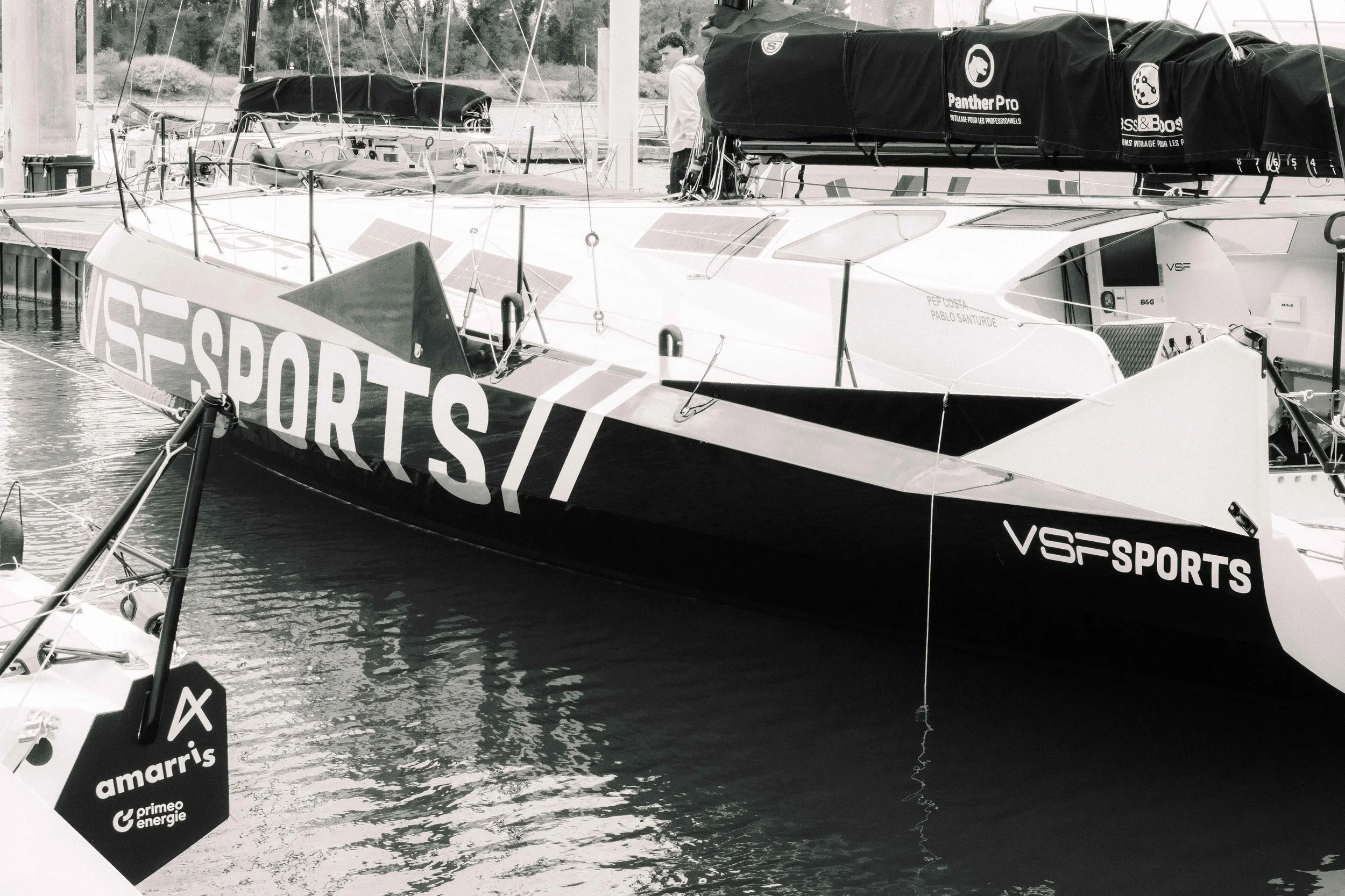 Yacht de sport amarré au port avec le logo 'VSF SPORTS' visible sur la coque, et un homme debout à l'arrière, vu en blanc et noir.