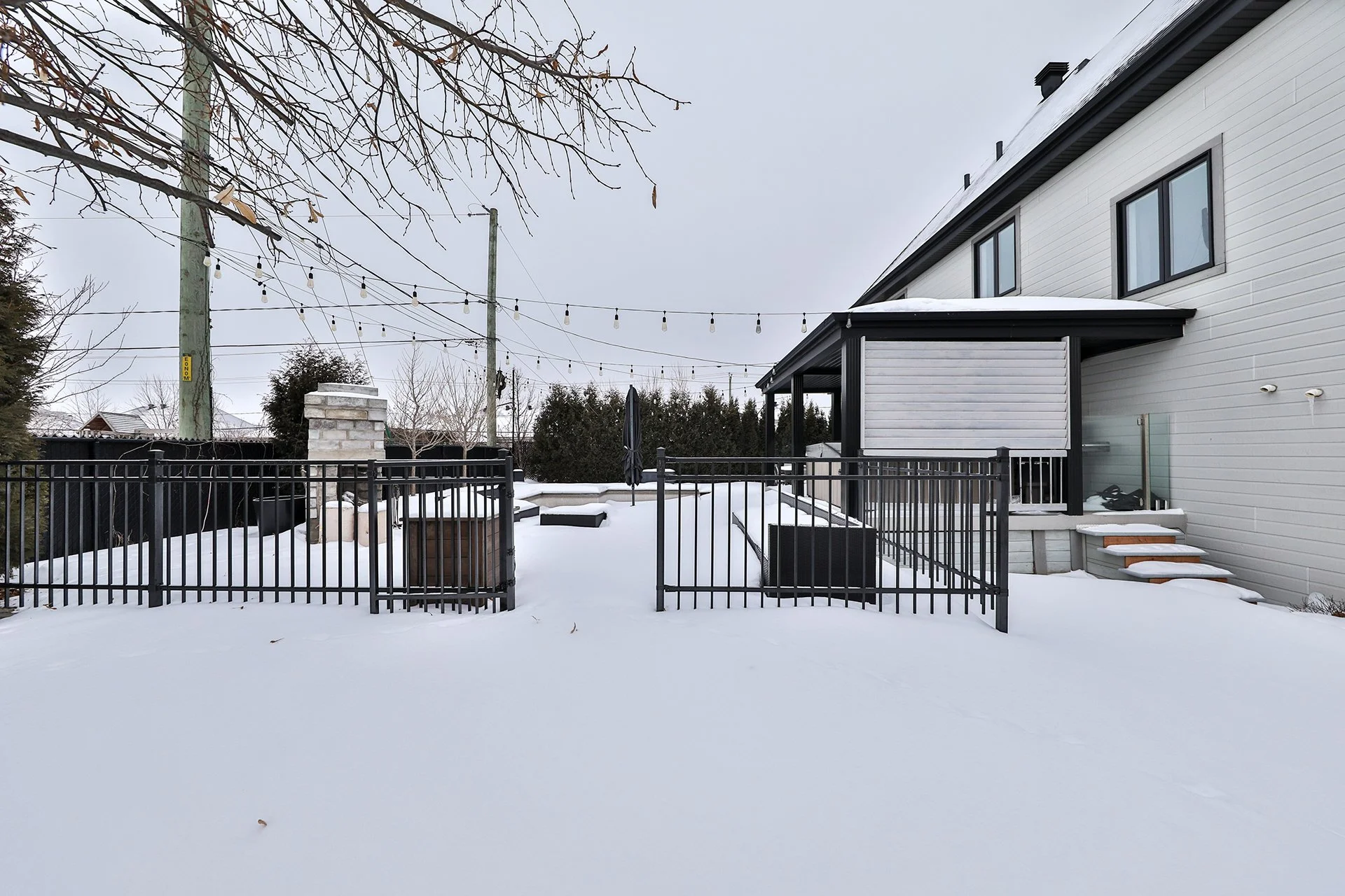 Une arrière-cour enneigée avec une clôture noire, une terrasse, une maison moderne blanche avec un porche, des escaliers en bois recouverts de neige, un arbre sans feuilles, des câbles électriques suspendus, un ciel gris.