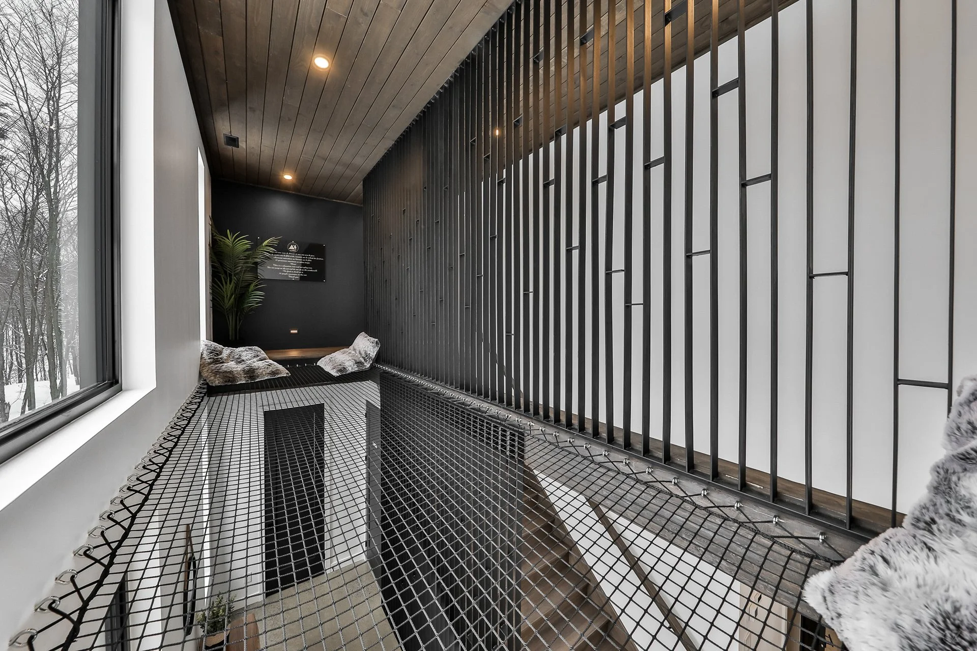 Balcon en intérieur avec un mur en métal noir, un plafond en bois et un filet de sécurité en métal, avec des coussins en fausse fourrure et une plante verte