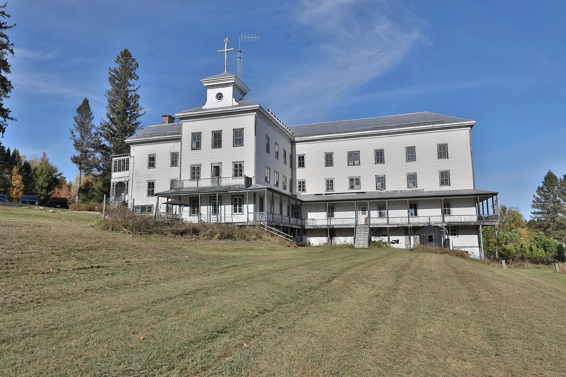 Grand bâtiment blanc en dehors, avec plusieurs fenêtres, balcon, et tours. Sur le toit, croix et antennes. Environnement avec arbres et ciel bleu.