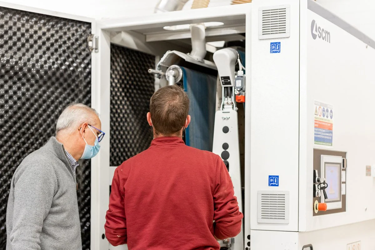 Deux hommes regardent une machine technique ou un appareil industriel dans une salle avec des murs isolants. L'un porte un masque facial, l'autre a les cheveux courts et porte un pull rouge.