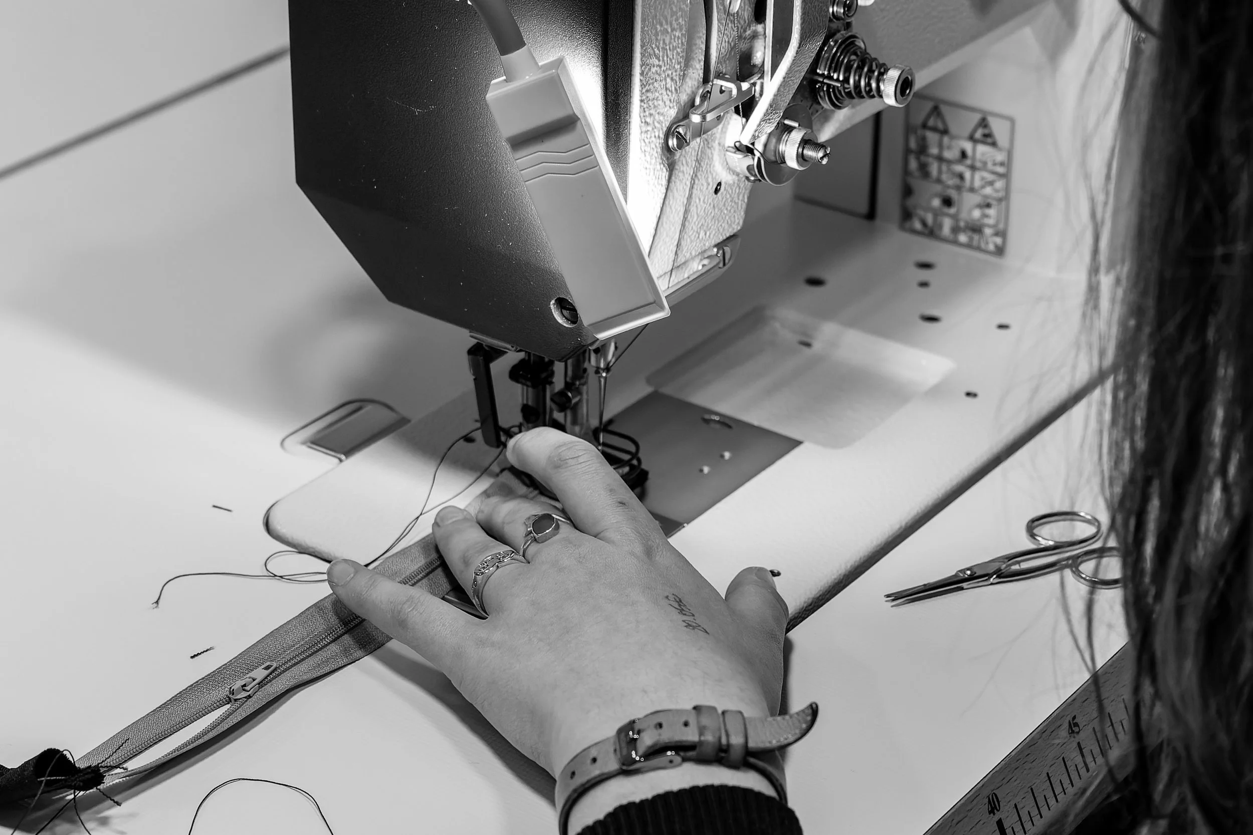 Une personne cousant une fermeture éclair avec une machine à coudre. La personne porte une bague et un bracelet, avec des ciseaux à proximité.