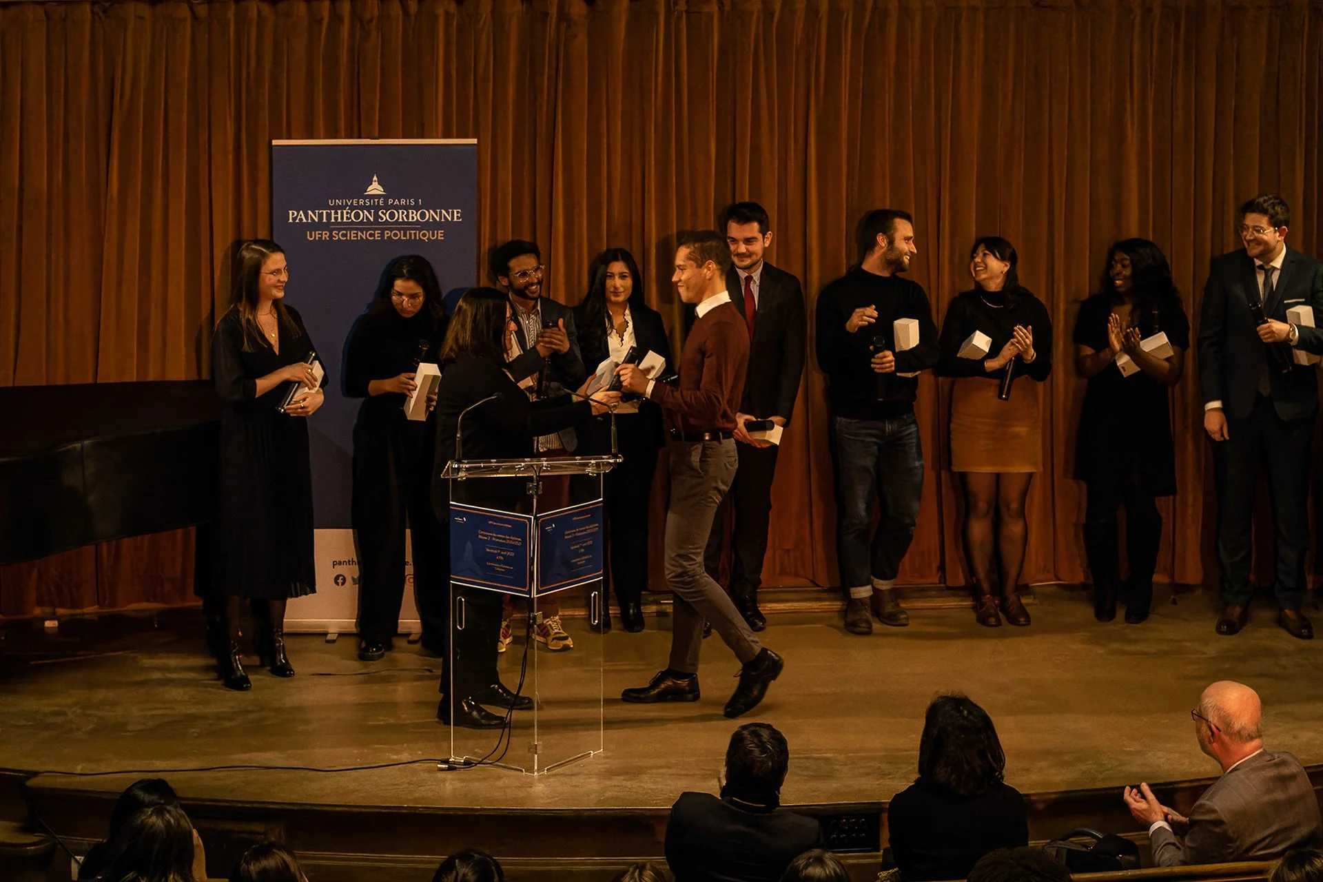 Cérémonie académique avec un groupe de jeunes personnes sur scène recevant des prix, avec un panneau en arrière-plan indiquant l'université Paris 1 Panthéon Sorbonne.