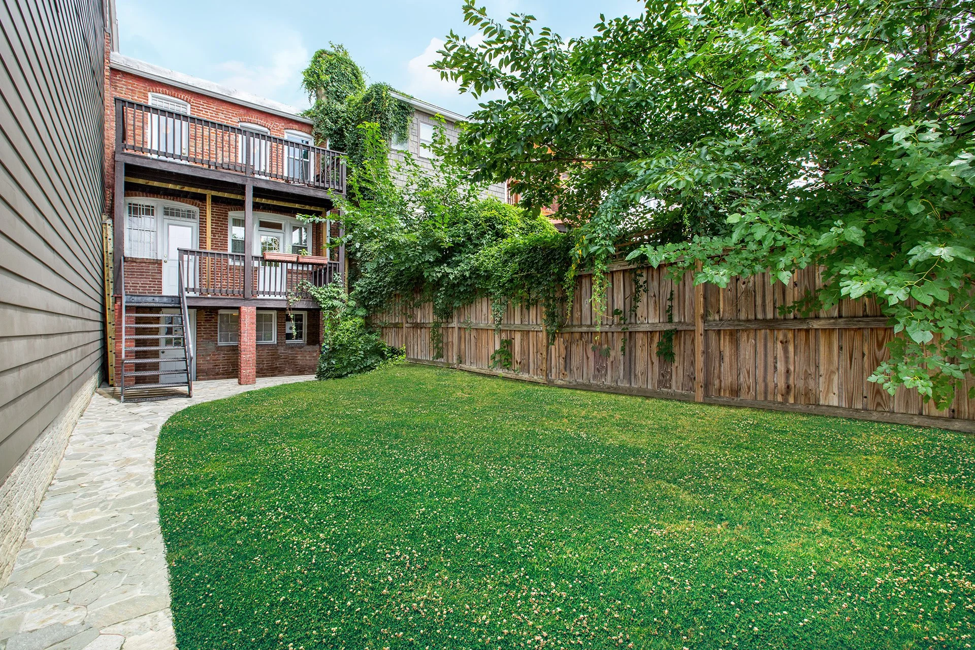 Jardin privé avec pelouse verte, clôture en bois, arbres, balcon en arrière-plan d'un bâtiment en briques rouges.