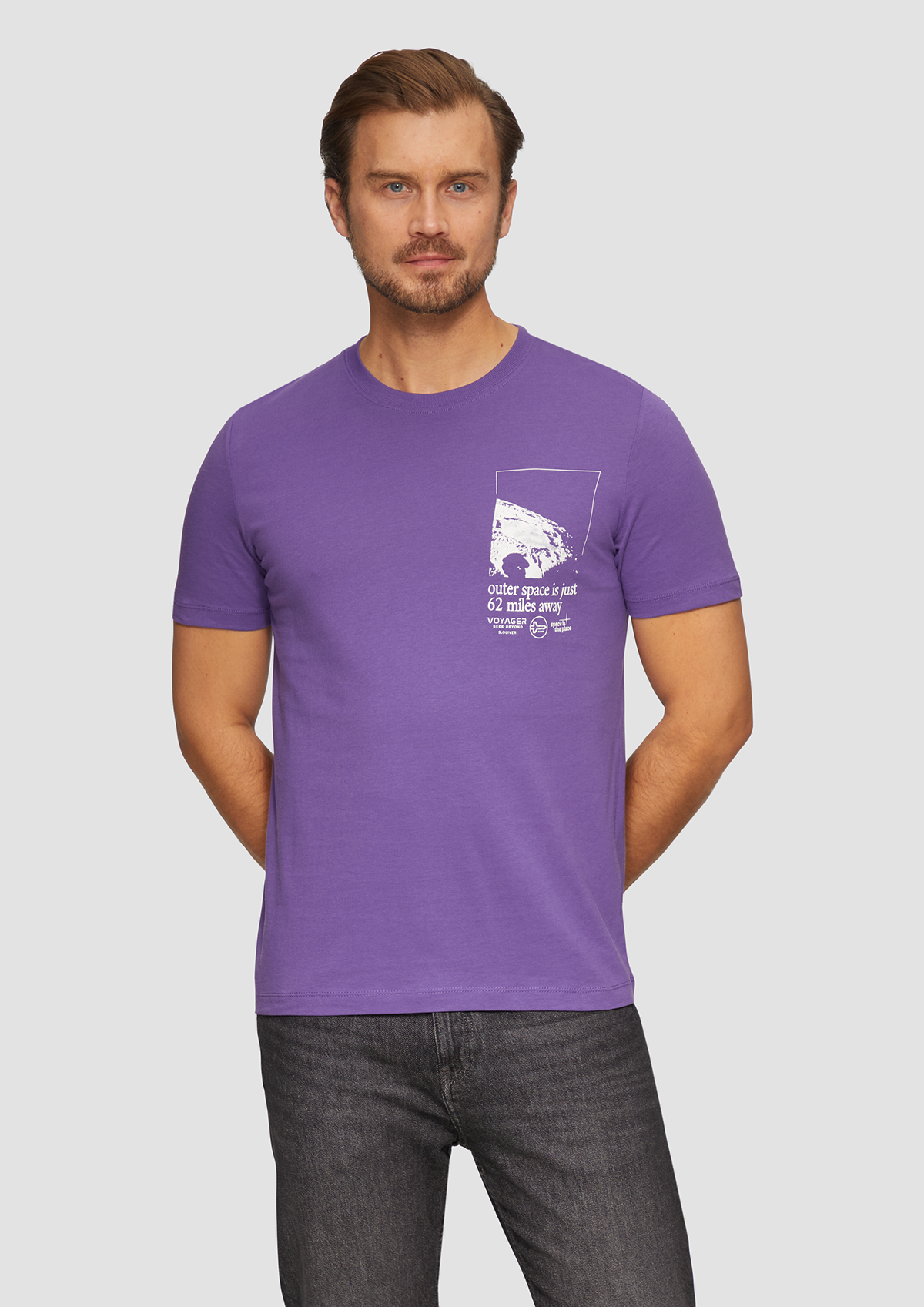 Homme portant un t-shirt violet avec impression blanche, debout face à un fond gris clair.