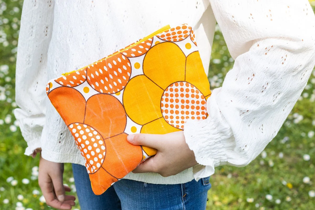 Personne tenant un sac de tissu orange avec un motif de cercles de différentes tailles et couleurs, portant un haut blanc à manches longues et un pantalon en jean, dans un extérieur avec de l'herbe et des fleurs.