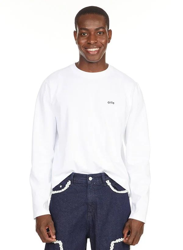 Une personne souriante porte un sweat blanc portant le mot 'arte' et un pantalon bleu foncé avec des détails perlés blancs.