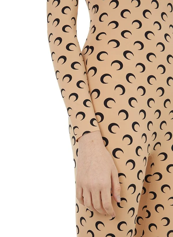 Partie d'une personne en combinaison beige à motifs de croissants noirs, avec la main reposant sur la cuisse, fond blanc.