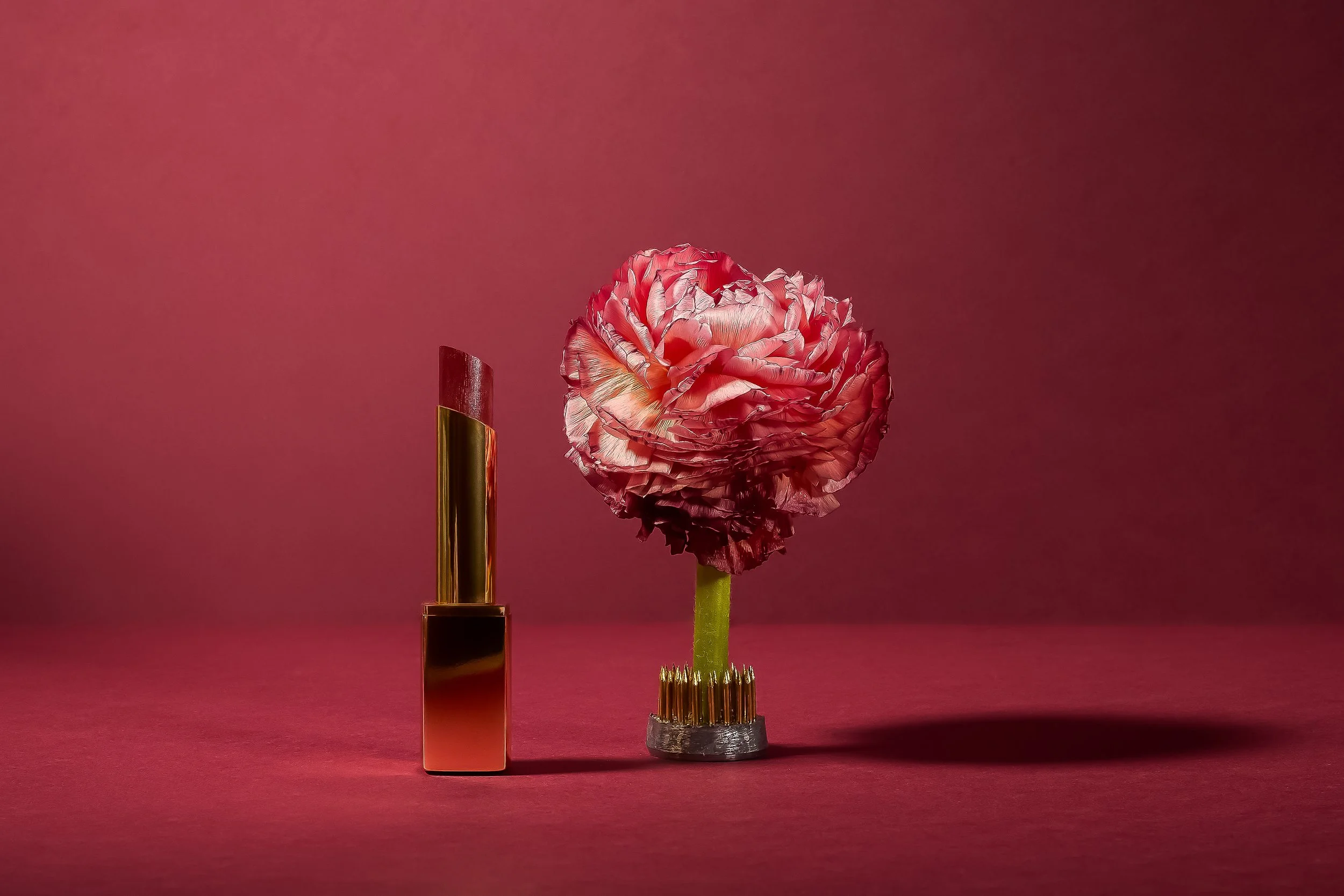 Un rouge à lèvres doré et un porte-bouquet en forme de bouchon contenant une fleur rose, tous deux placés sur un fond rouge.