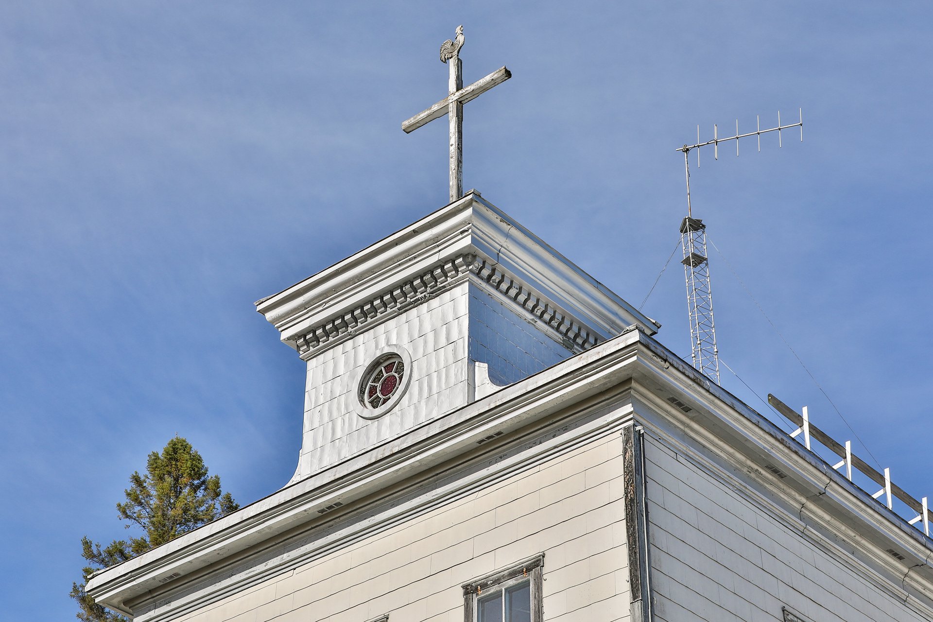 Le toit d'un bâtiment blanc avec une croix en haut et une petite fenêtre. Le ciel est bleu avec quelques nuages et un arbre est visible à gauche.