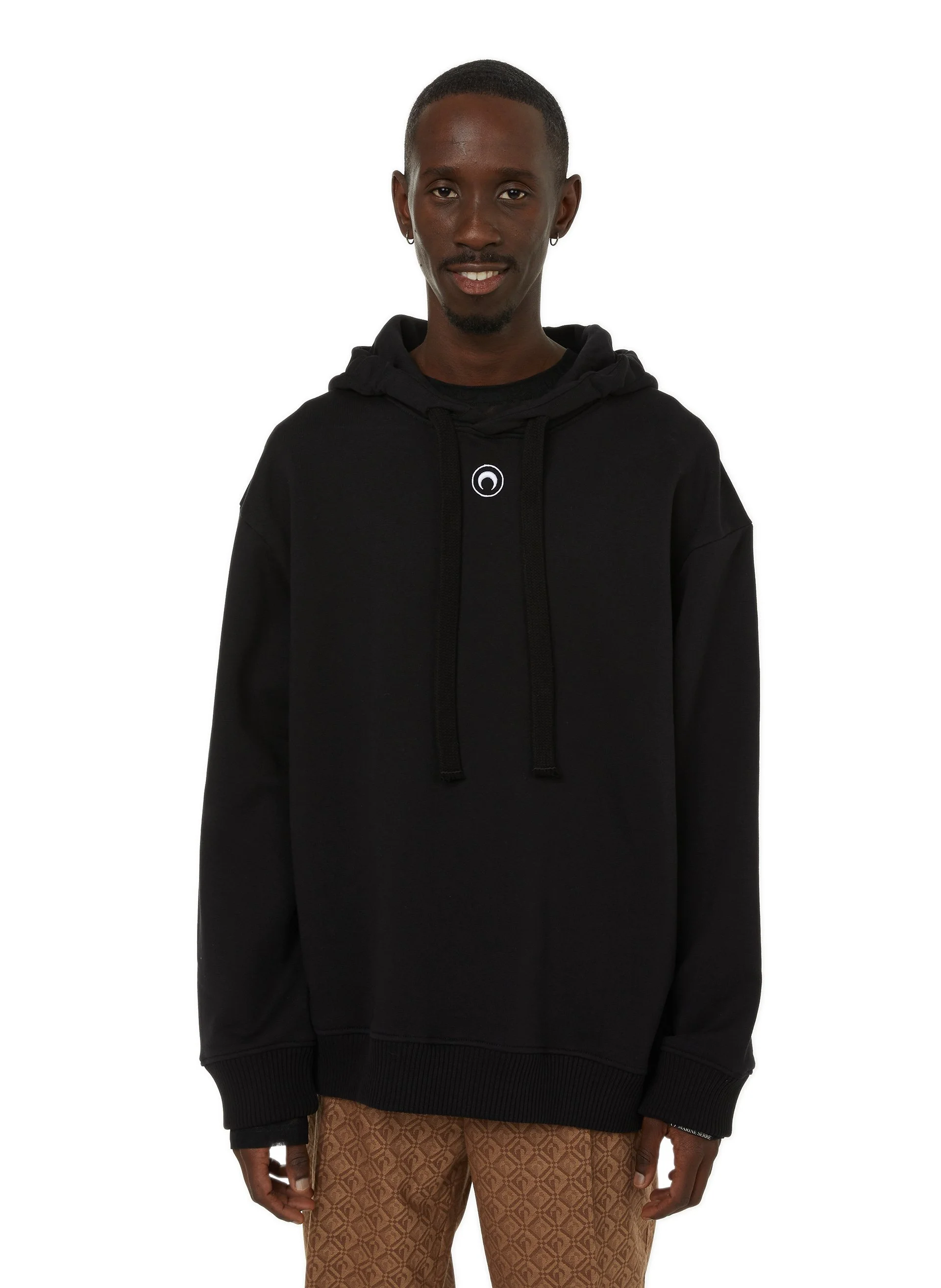 Un homme souriant porte un sweat à capuche noir avec un logo blanc au centre, et un pantalon marron avec un motif géométrique.