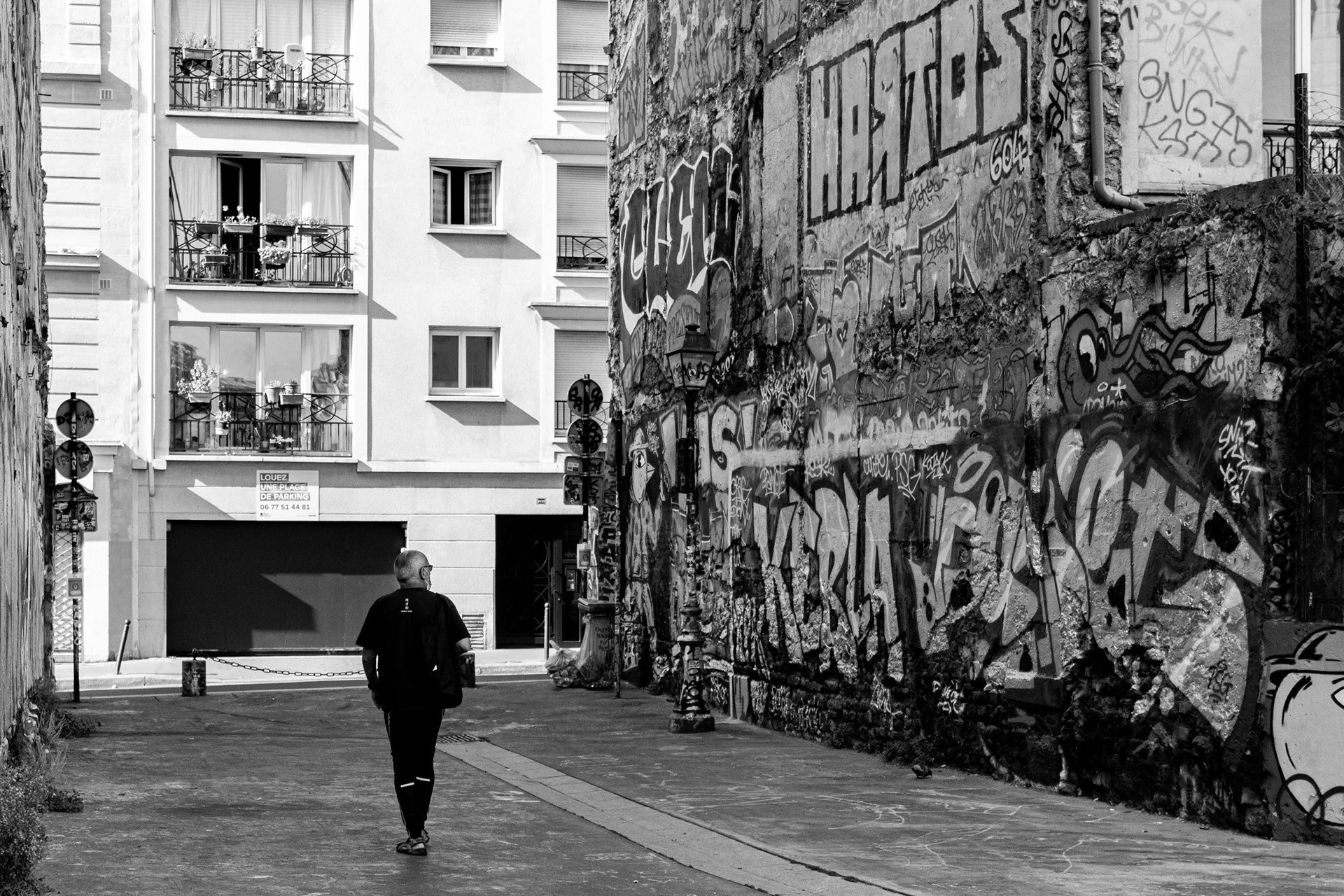 Un homme marchant seul dans une ruelle urbaine entre un mur de graffiti et un bâtiment résidentiel.