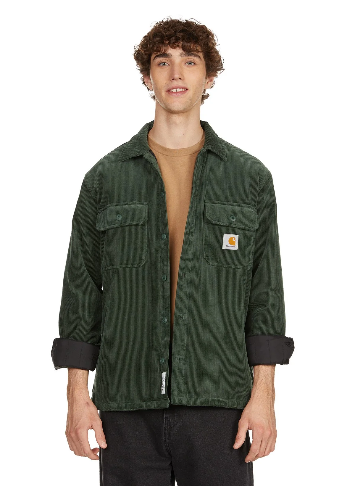 Un jeune homme porte une chemise en velours côtelé vert avec une étiquette Carhartt, chemise ouverte sur un t-shirt beige, fond blanc.