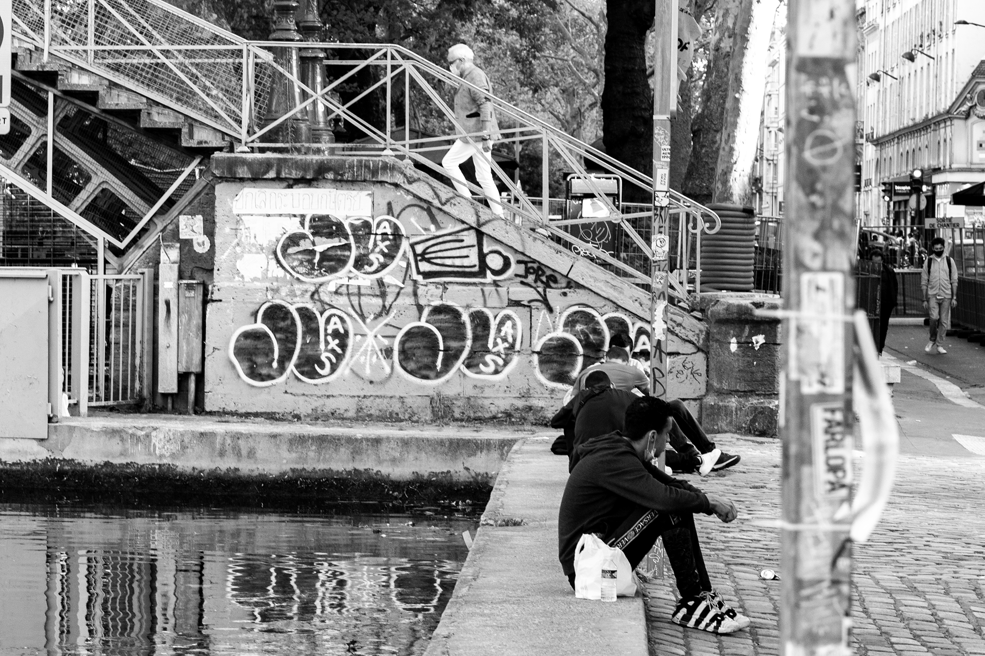 Quatre jeunes hommes assis au bord de l'eau sur un quai en pierre, deux autres personnes marchent en haut des escaliers en arrière-plan, une personne portant un masque dans une rue urbaine. Graffiti sur un mur en face.