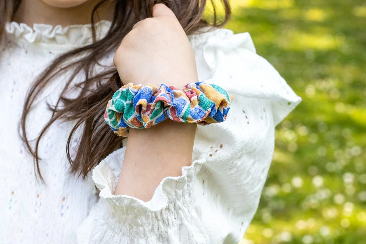 Poignet d'une femme portant un bracelet en tissu multicolore, avec un vêtement blanc en dentelle, à l'extérieur avec de la verdure en arrière-plan.