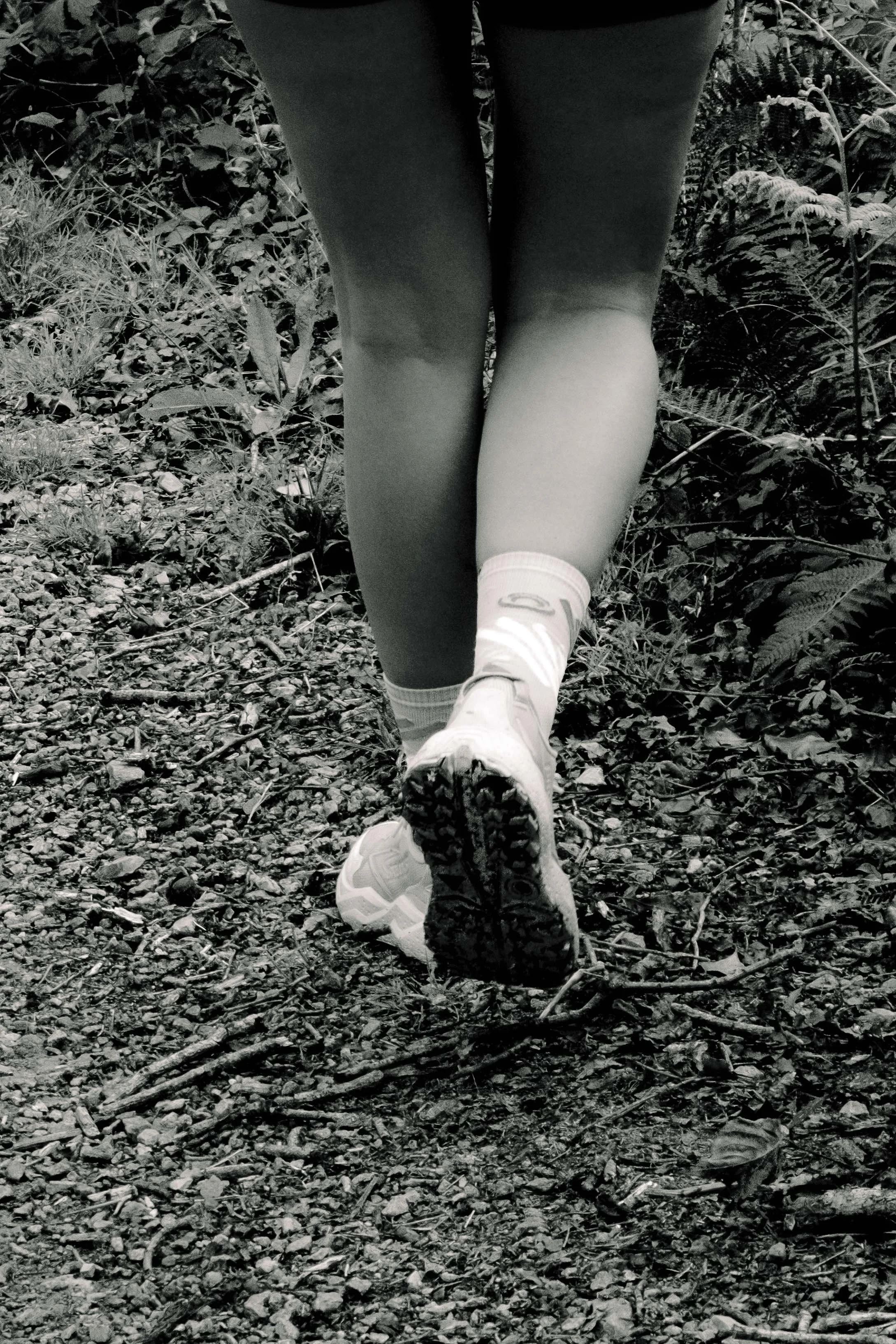 Jambes et chaussures d'une personne marchant sur un sentier en forêt, en noir et blanc.