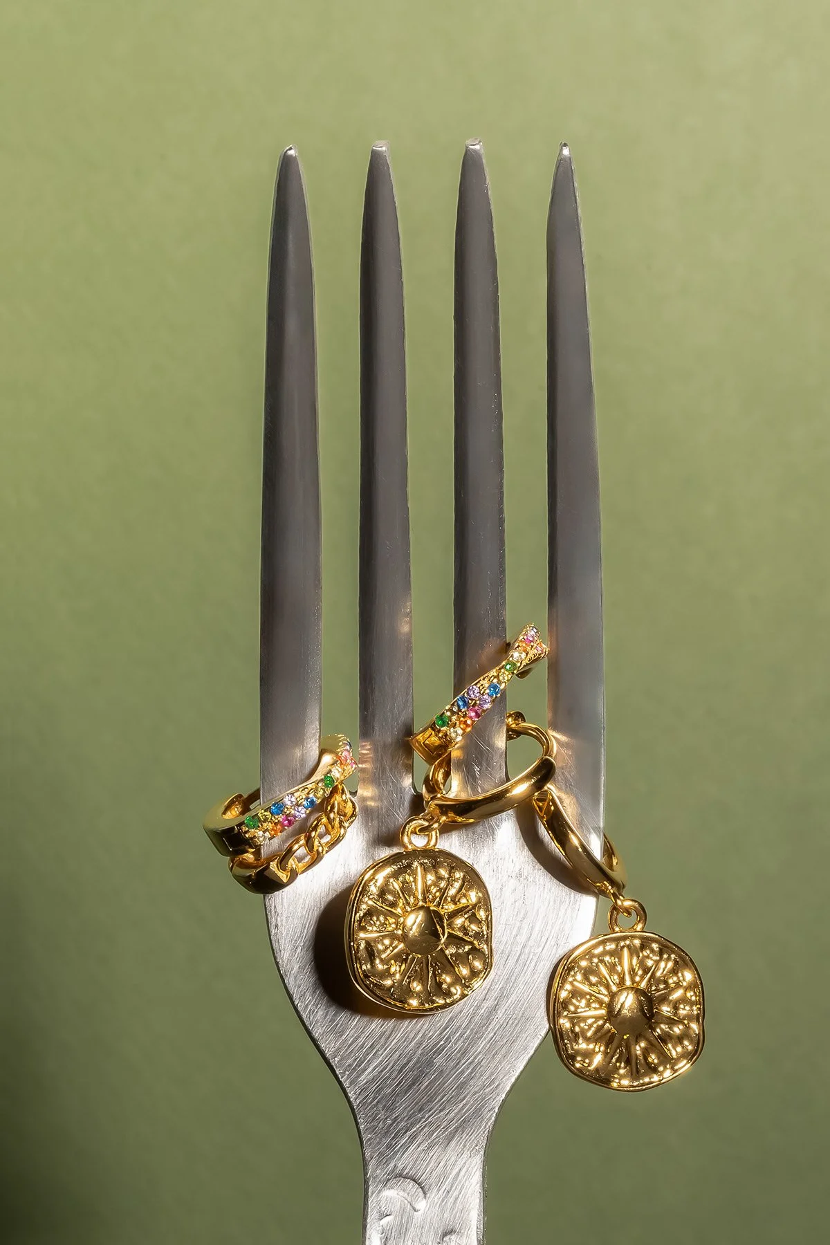 Bijoux dorés avec pierres colorées, posés sur une fourchette en acier inoxydable contre un fond vert.