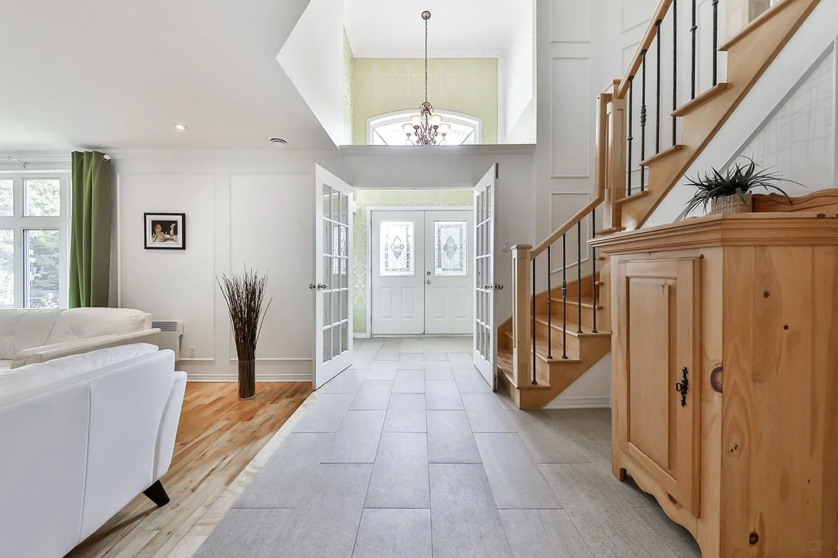 Intérieur d'une maison lumineuse avec porche en entrant, porte d'entrée double en verre, escalier en bois avec balustrade en métal, mobilier en bois et décoration intérieure.