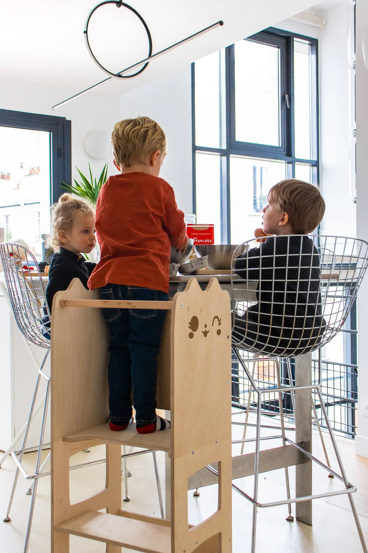 Trois enfants jouent ou discutent autour d'une table dans une cuisine lumineuse. Deux sont assis sur des chaises en filaire et un debout sur un marchepied en bois avec un visage d'animal dessiné. La pièce est bien éclairée par de grandes fenêtres.