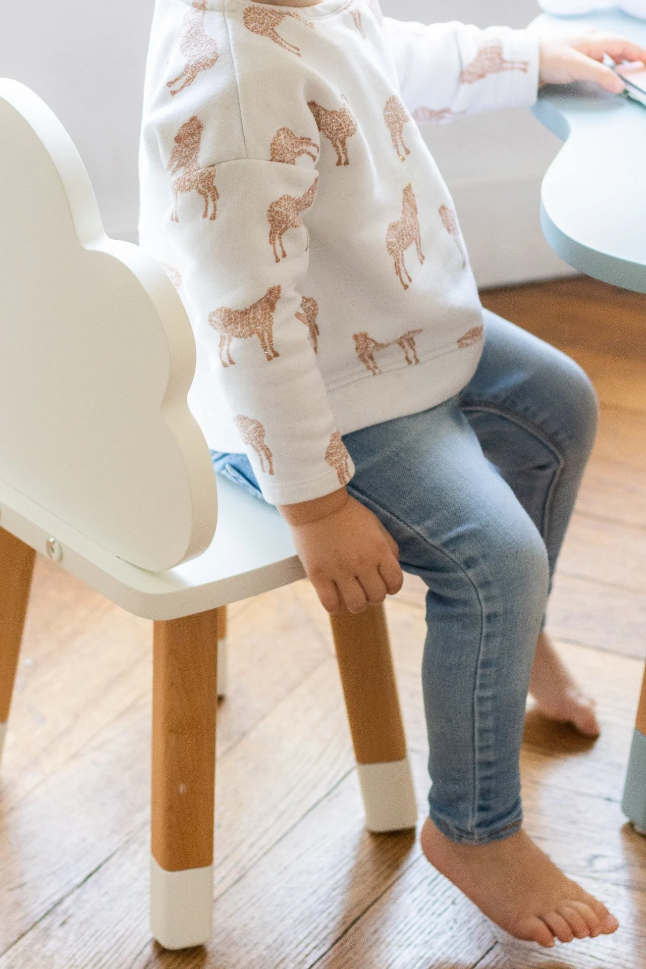 Enfant assis sur une chaise en bois, portant un sweat avec des illustrations de girafes, et des jeans, pieds nus, dans une pièce avec un sol en bois clair.