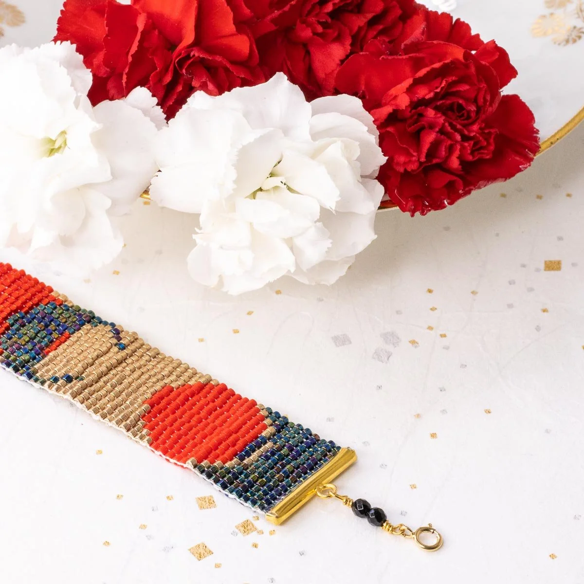 Un arrangement floral avec des fleurs rouges et blanches, une table blanche, décoration en confettis dorés, et un bracelet en perles multicolores