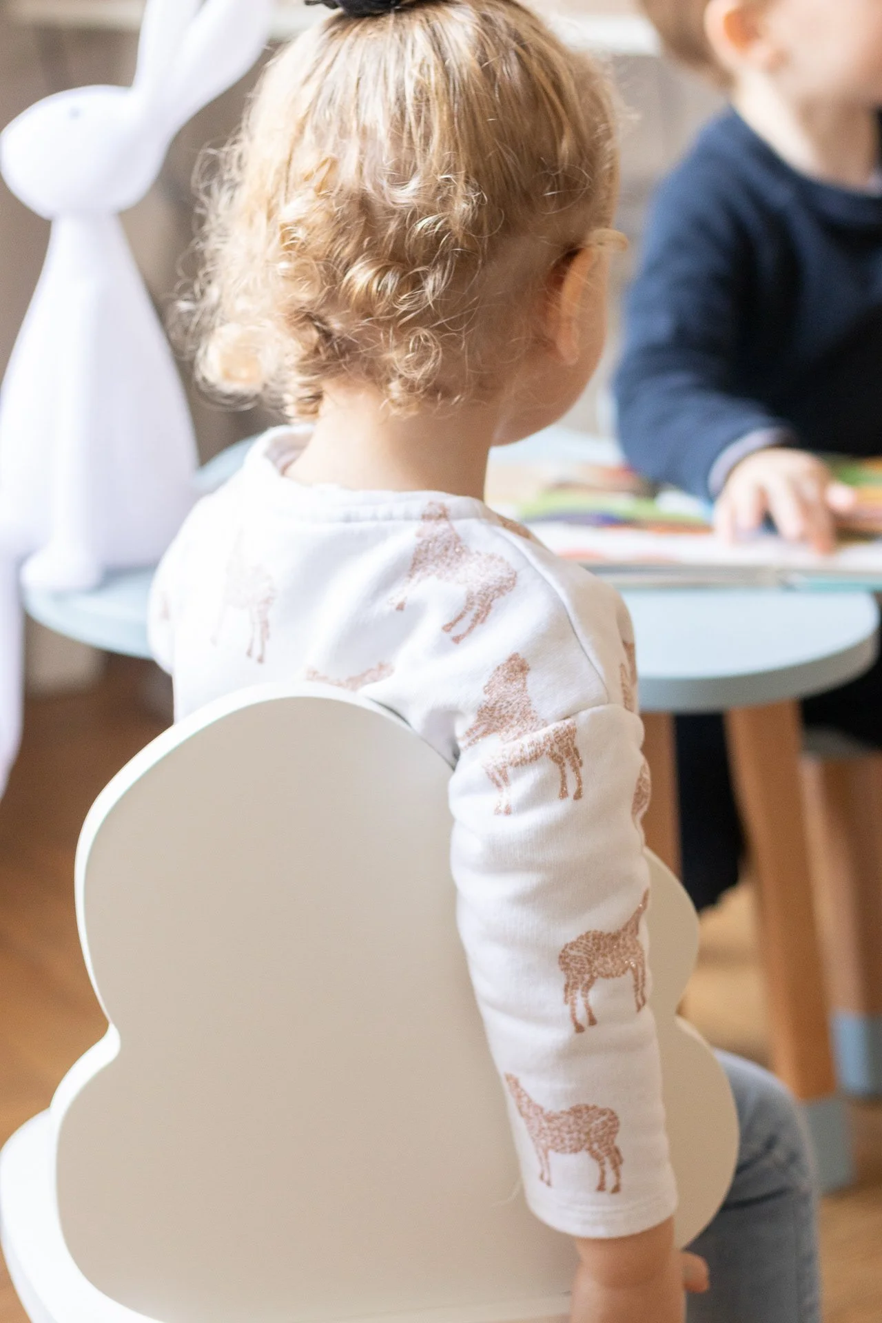 Enfant avec des cheveux bouclés roux assis sur une chaise en forme de nuage en bois, regardant un adulte qui lit ou montre quelque chose devant lui.
