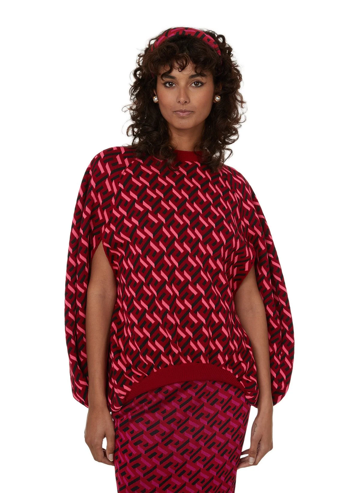 Une femme porte un ensemble à motif géométrique rouge, noir et rose, comprenant un haut à manches trapèzes et une jupe assortie, avec un bandeau coordonné sur la tête