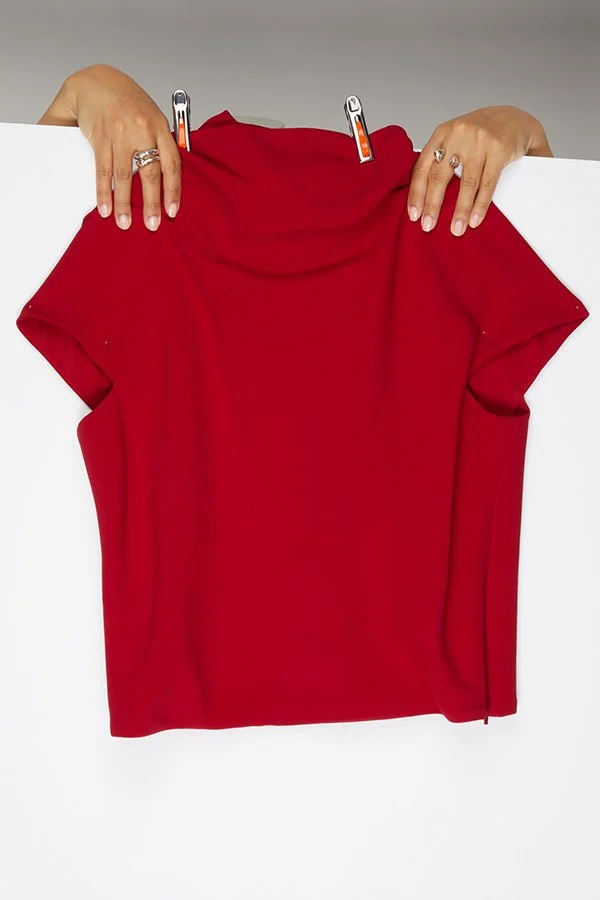 Deux mains tiennent une pièce de tissu rouge pliée. La pièce est suspendue par deux pinces clipsées à un support blanc.