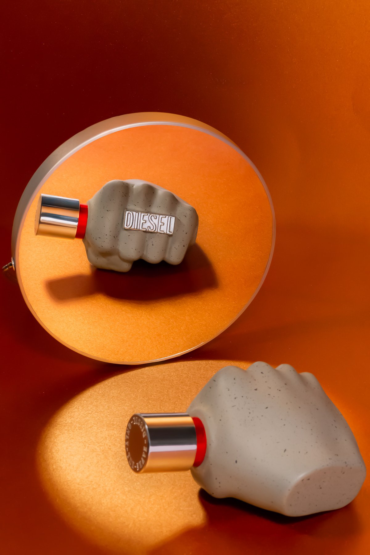 Deux bouchons de bouteille de parfum Dior en forme de poing, un sur une surface orange et l'autre dans un miroir, avec une lumière chaude.